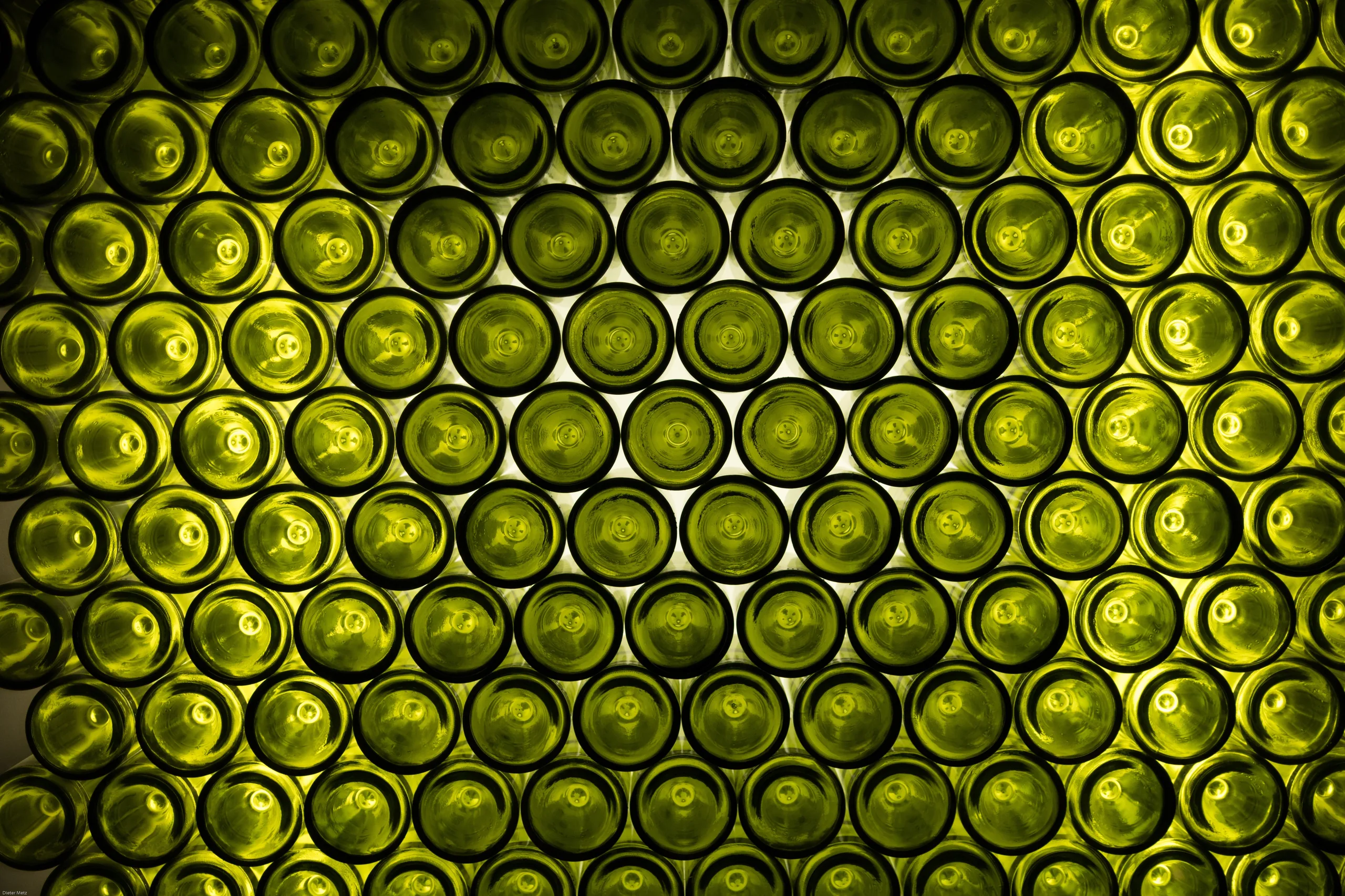 A wall of stacked wine bottles seen end-on, their green bases lit from behind