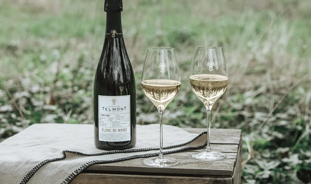 A bottle of Telmont Blanc de Noirs and two filled glasses on a wooden crate in a meadow