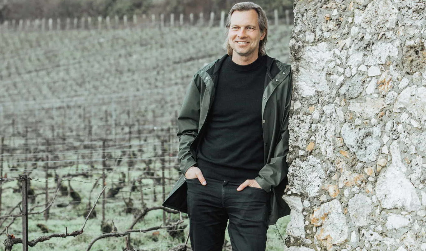A man in a green jacket leaning against a stone wall beside a winter vineyard