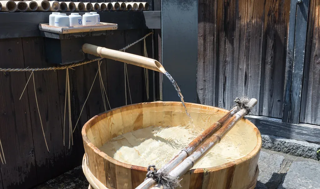 Spring water pouring from a bamboo spout into a cedar basin outside a Japanese brewery