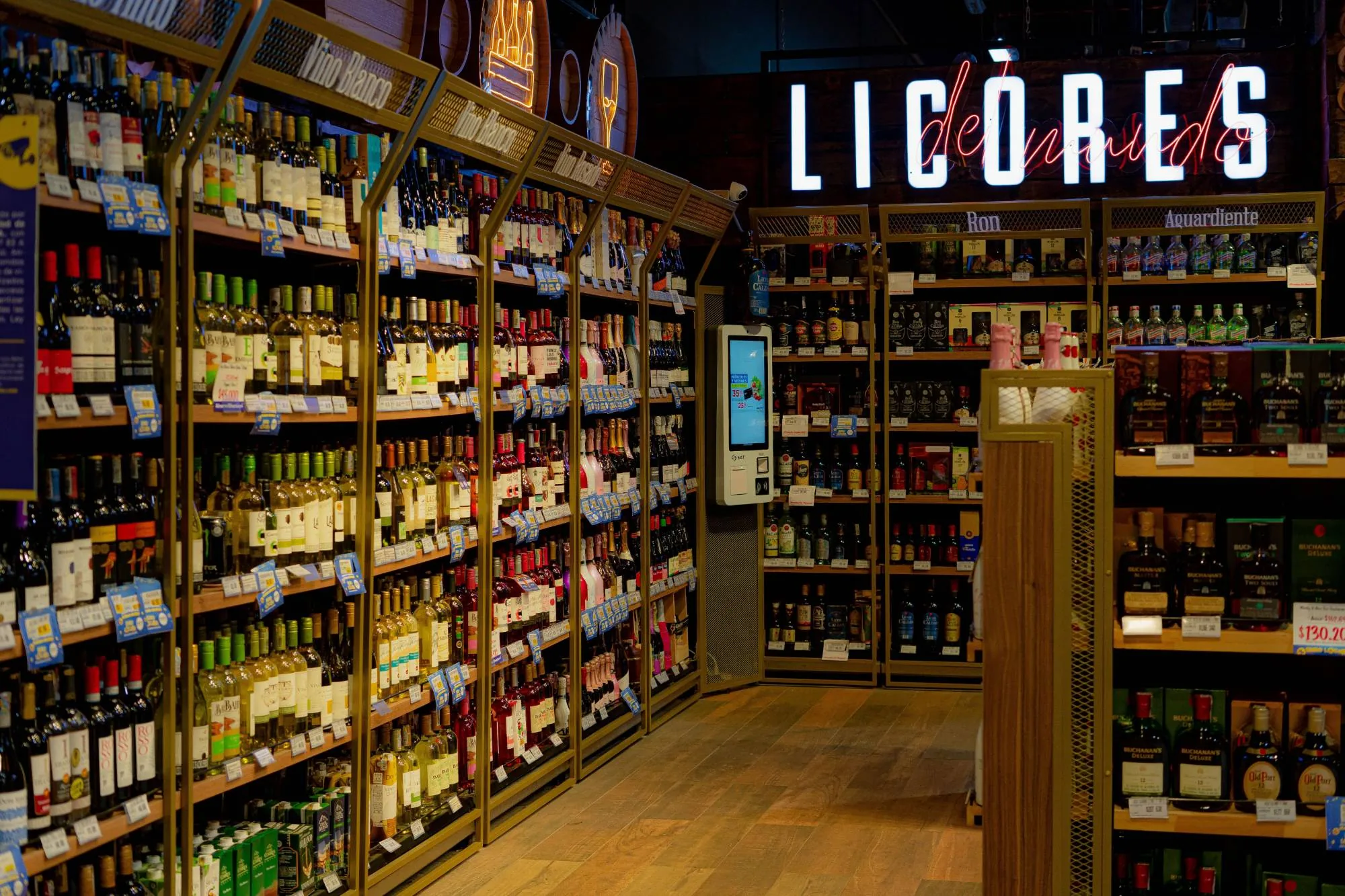 A liquor store aisle: wine shelves under Vino Blanco signs, a neon LICÒRES sign over the spirits beyond