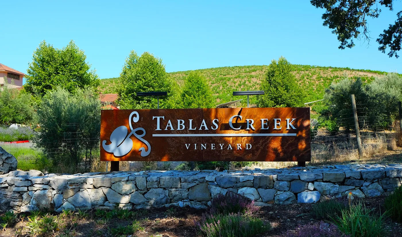 The corten-steel Tablas Creek Vineyard entrance sign on a dry-stone wall, with olive trees and a planted hillside behind