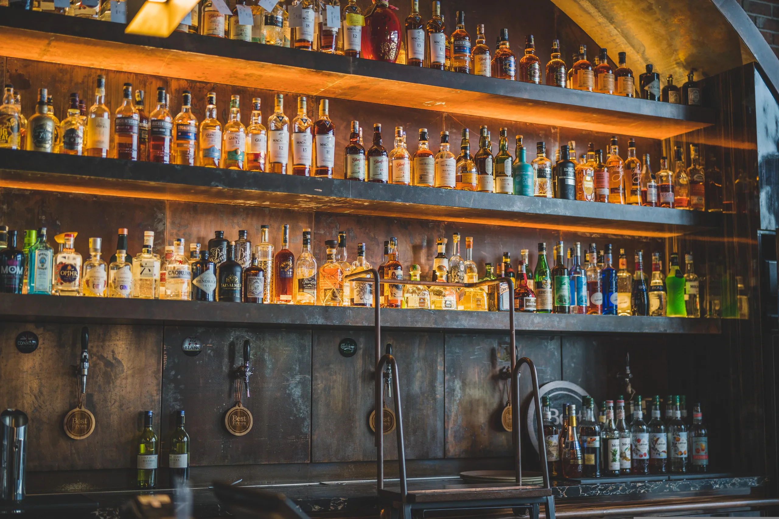 A bar back-shelf of spirits bottles lit warm above a run of beer taps