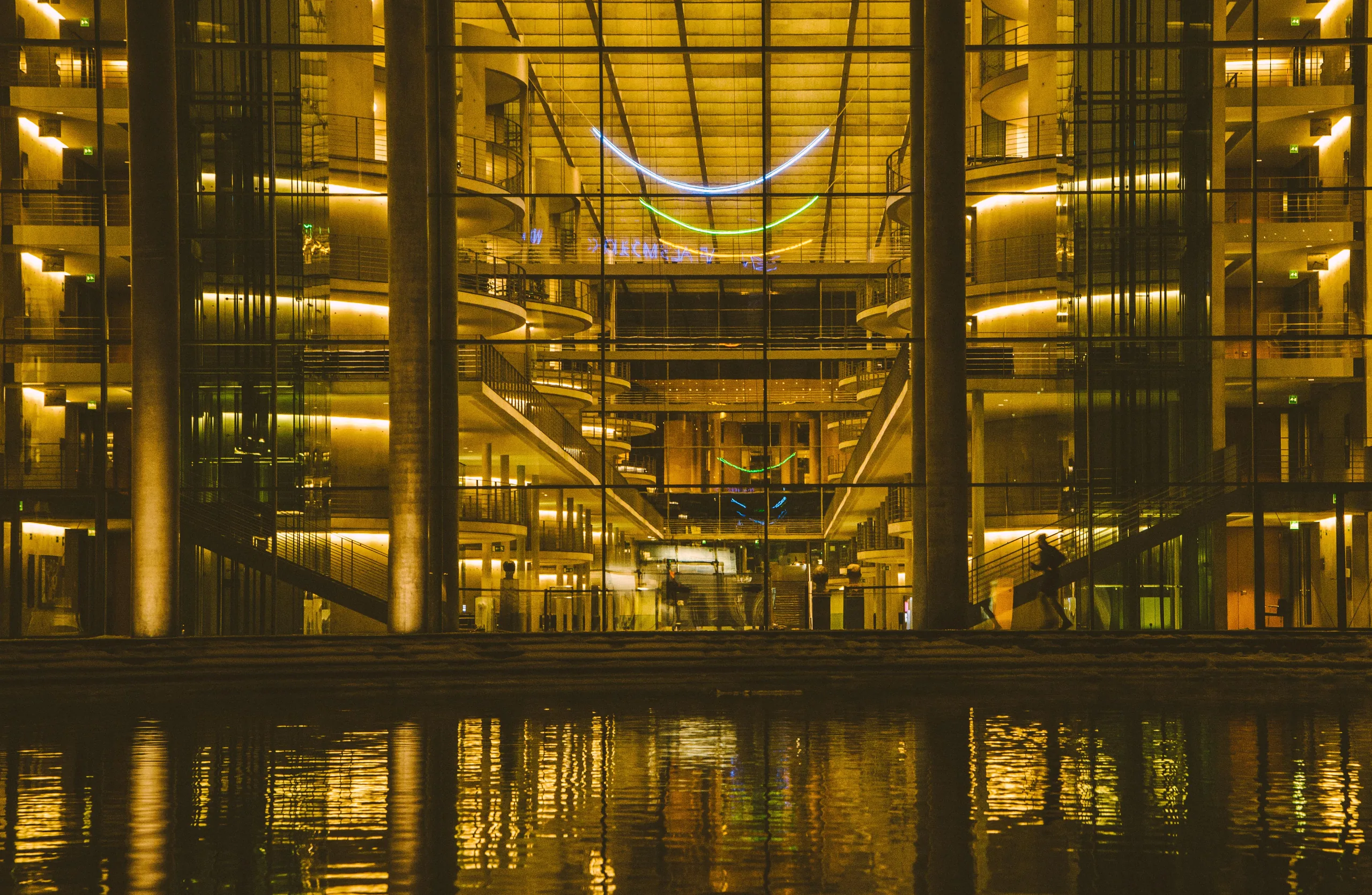 A glass-fronted building lit gold at night, its open floors and stairways reflected in the dark river in front of it