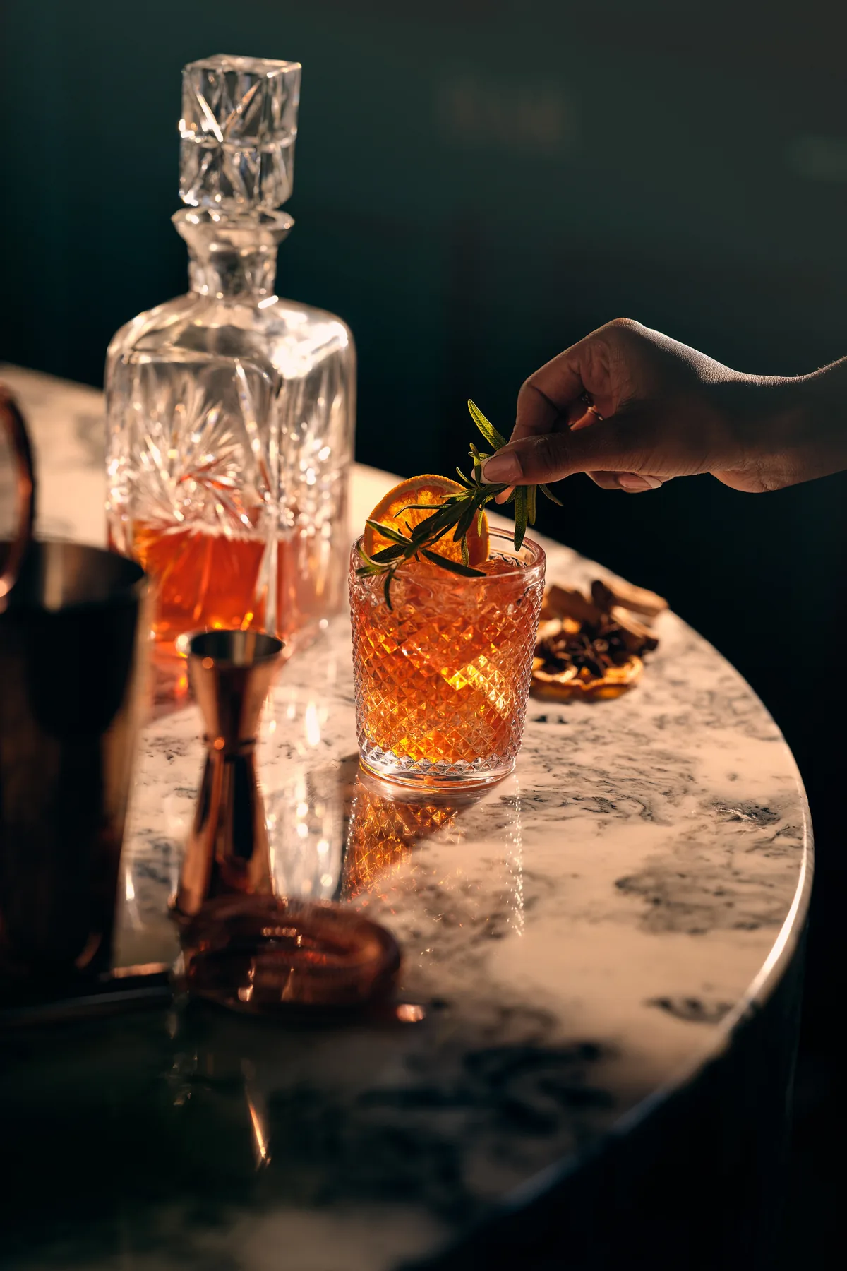 A hand placing a sprig of rosemary on an orange-garnished cocktail beside a cut-crystal decanter on a marble table
