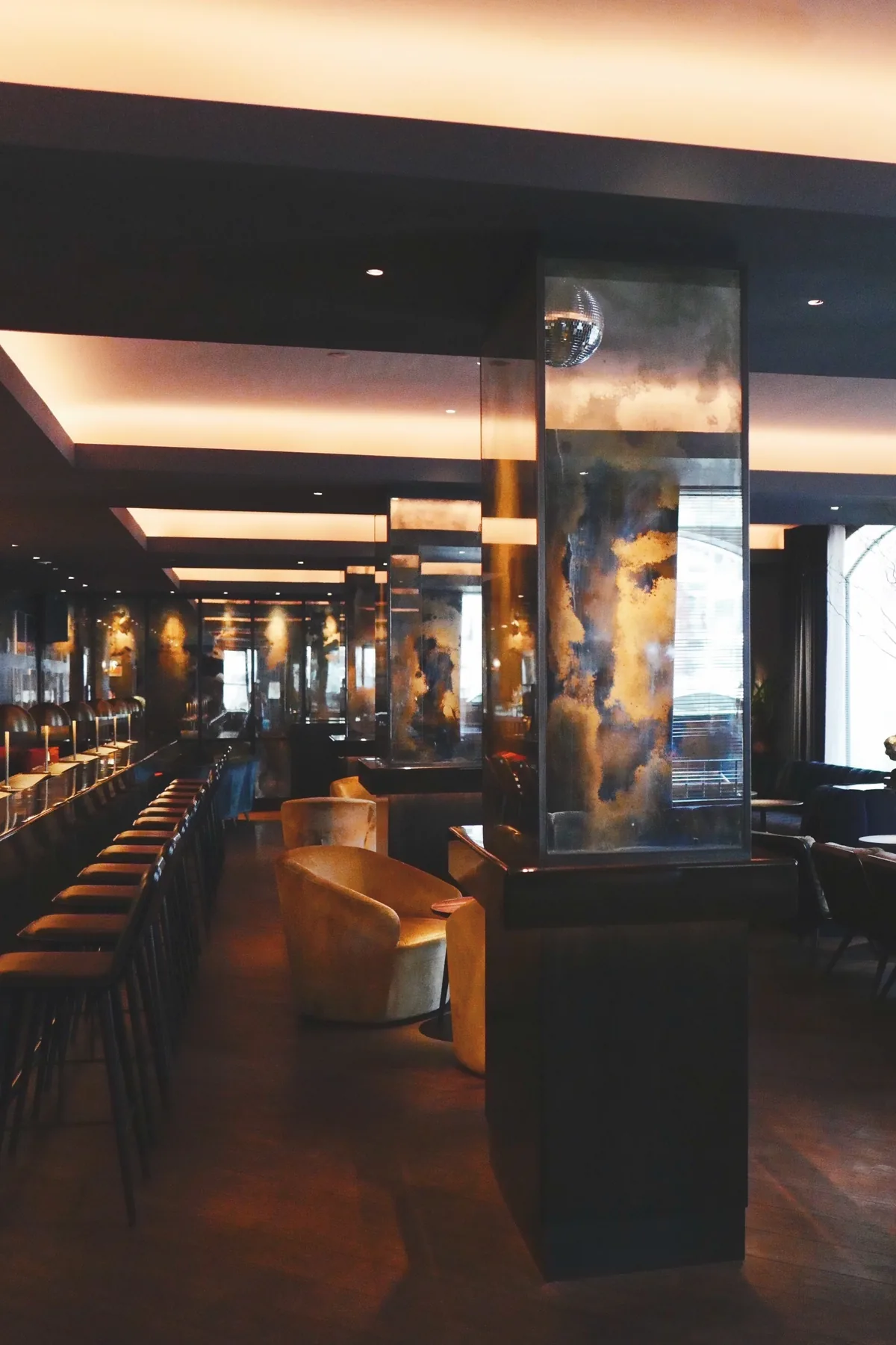 A long, dimly lit lounge bar with a row of bar stools, leather armchairs and a tall glass-cased column