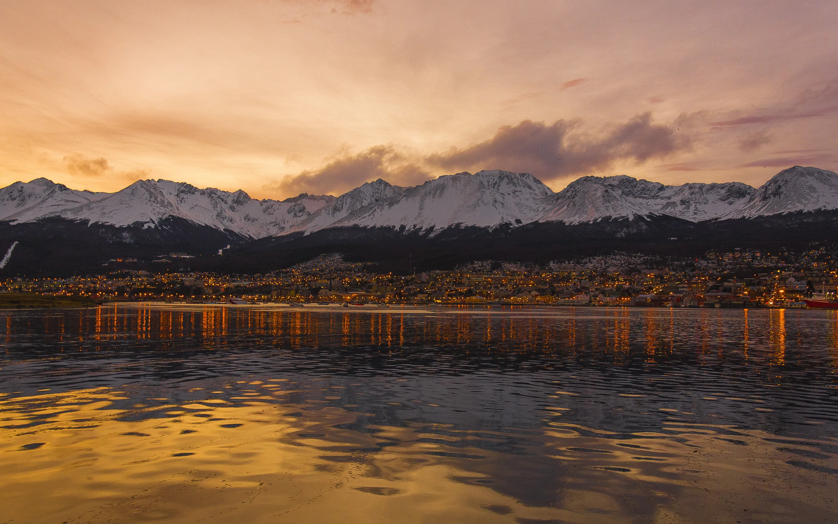 A town strung along a shoreline at dusk beneath a long range of snow-covered mountains, its lights doubled in still water under an orange sky.