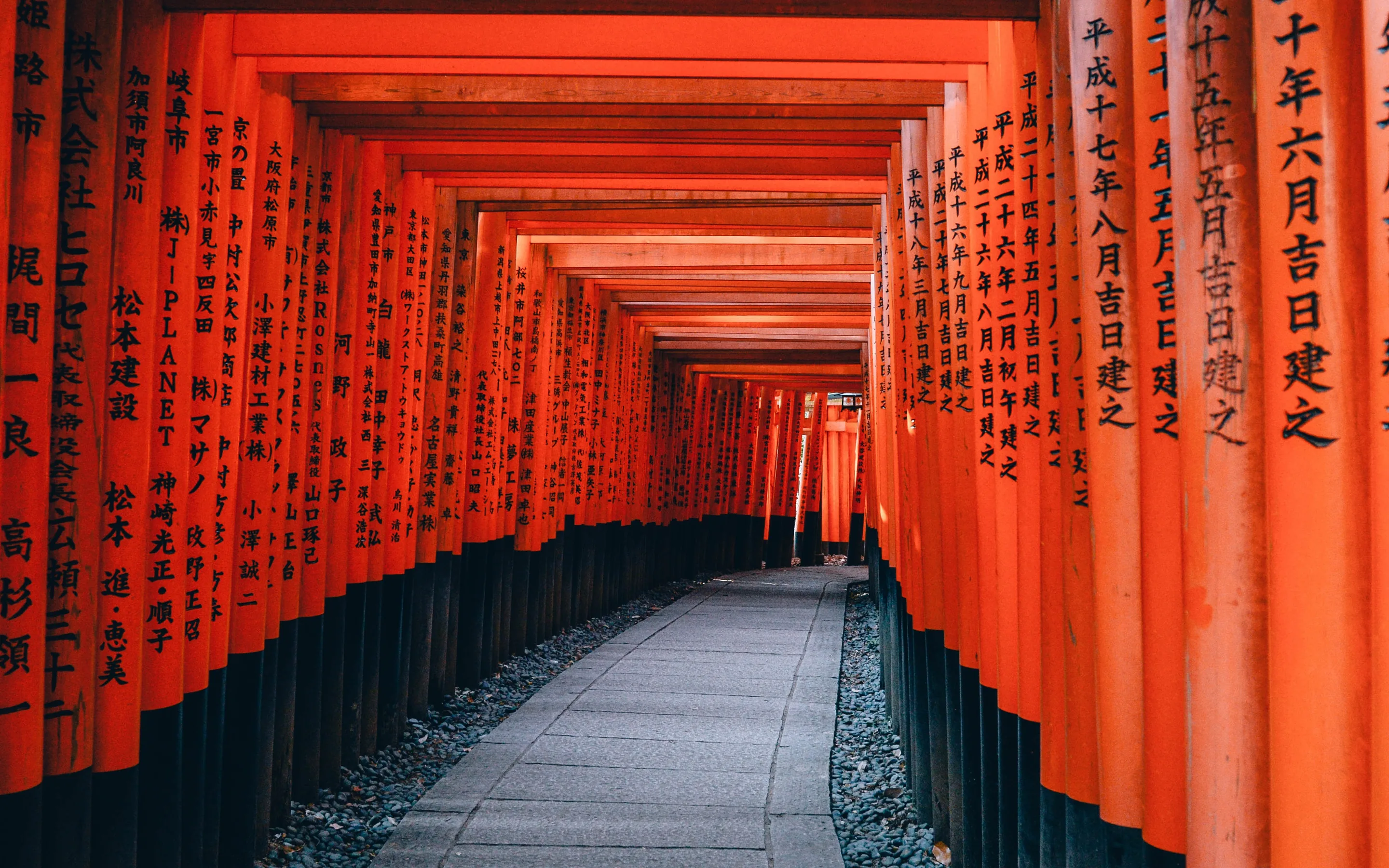 A tunnel of vermilion torii gates running downhill over a paved stone path, each upright inscribed with black characters