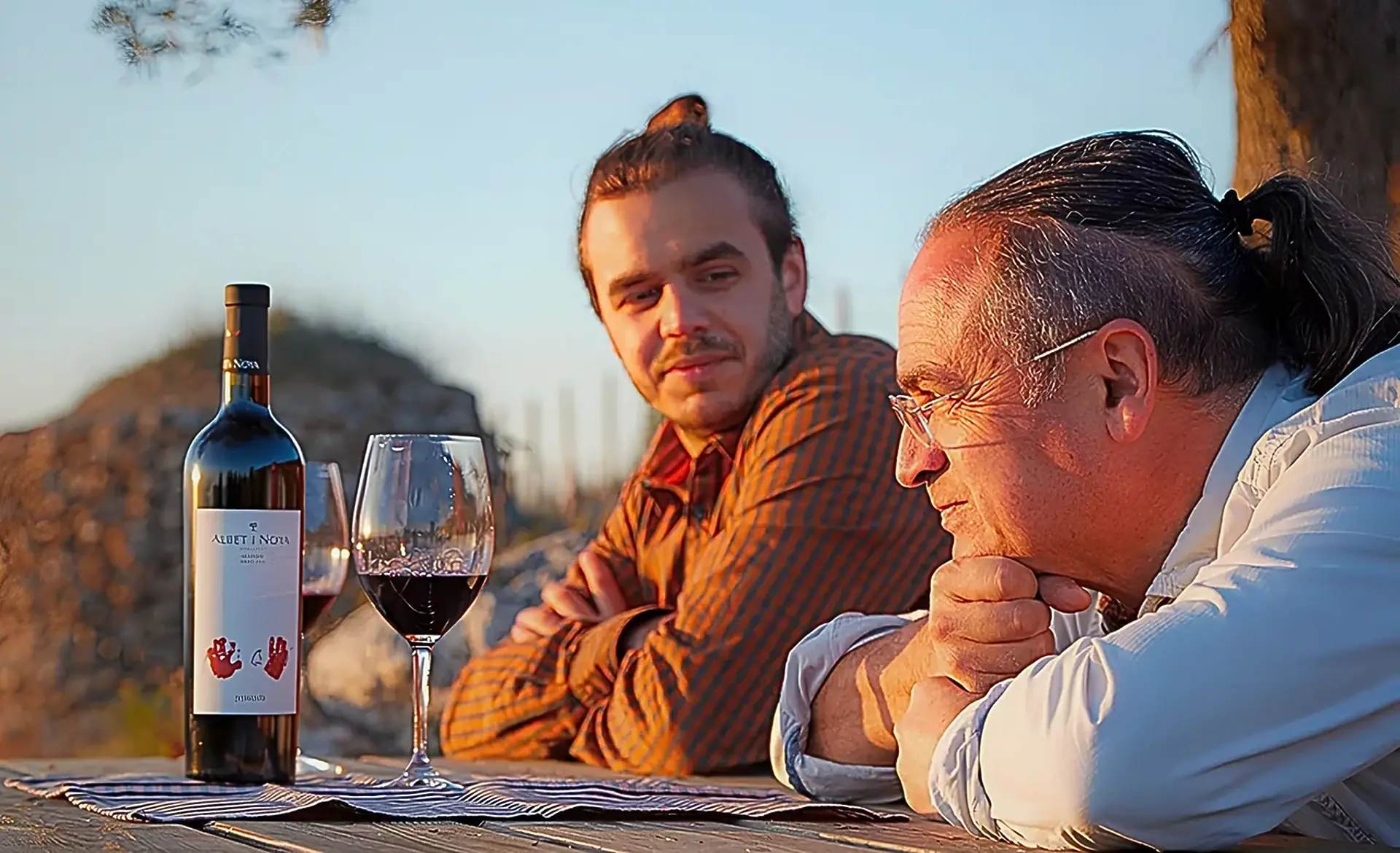 Two men at a wooden table at sunset with two glasses of red wine and a bottle labelled Albet i Noya