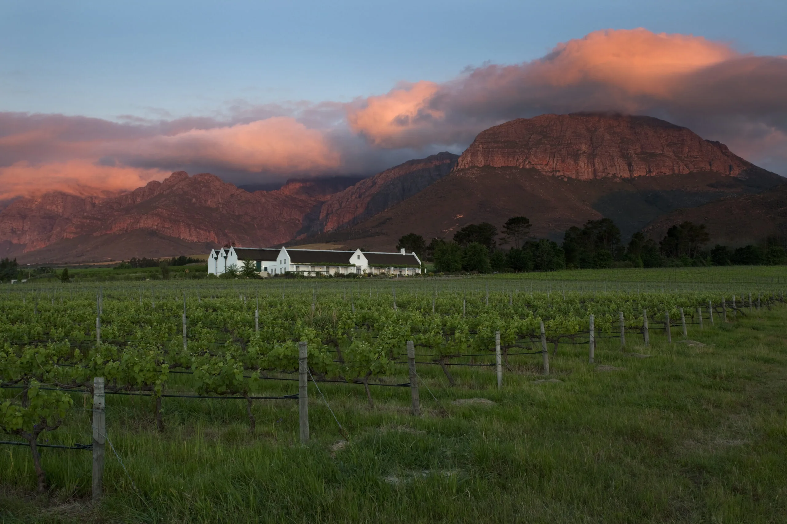 Whitewashed farmhouse at Avondale among vineyards beneath red rock peaks lit by sunset cloud