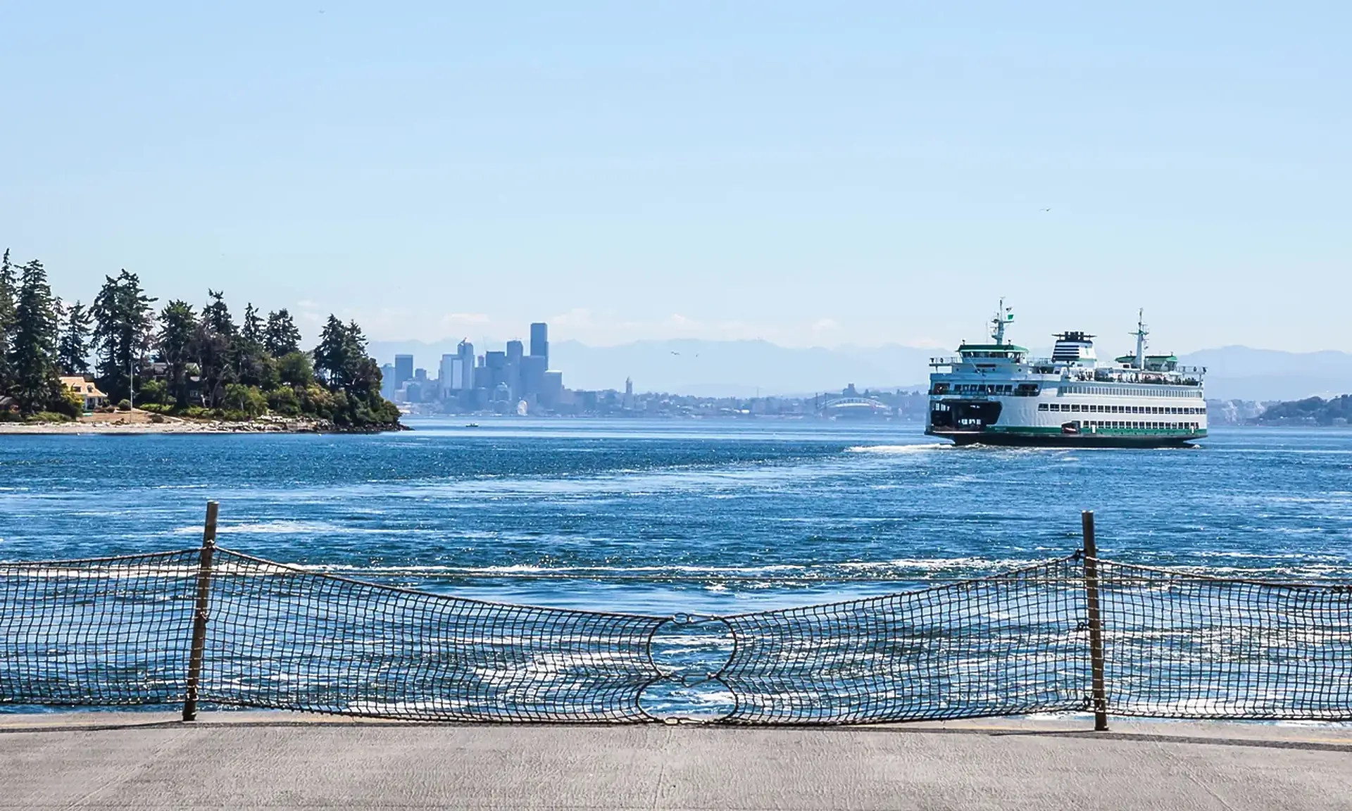 Green and white car ferry crossing choppy blue water toward a city skyline, a wooded point at left and a netted dock railing in front
