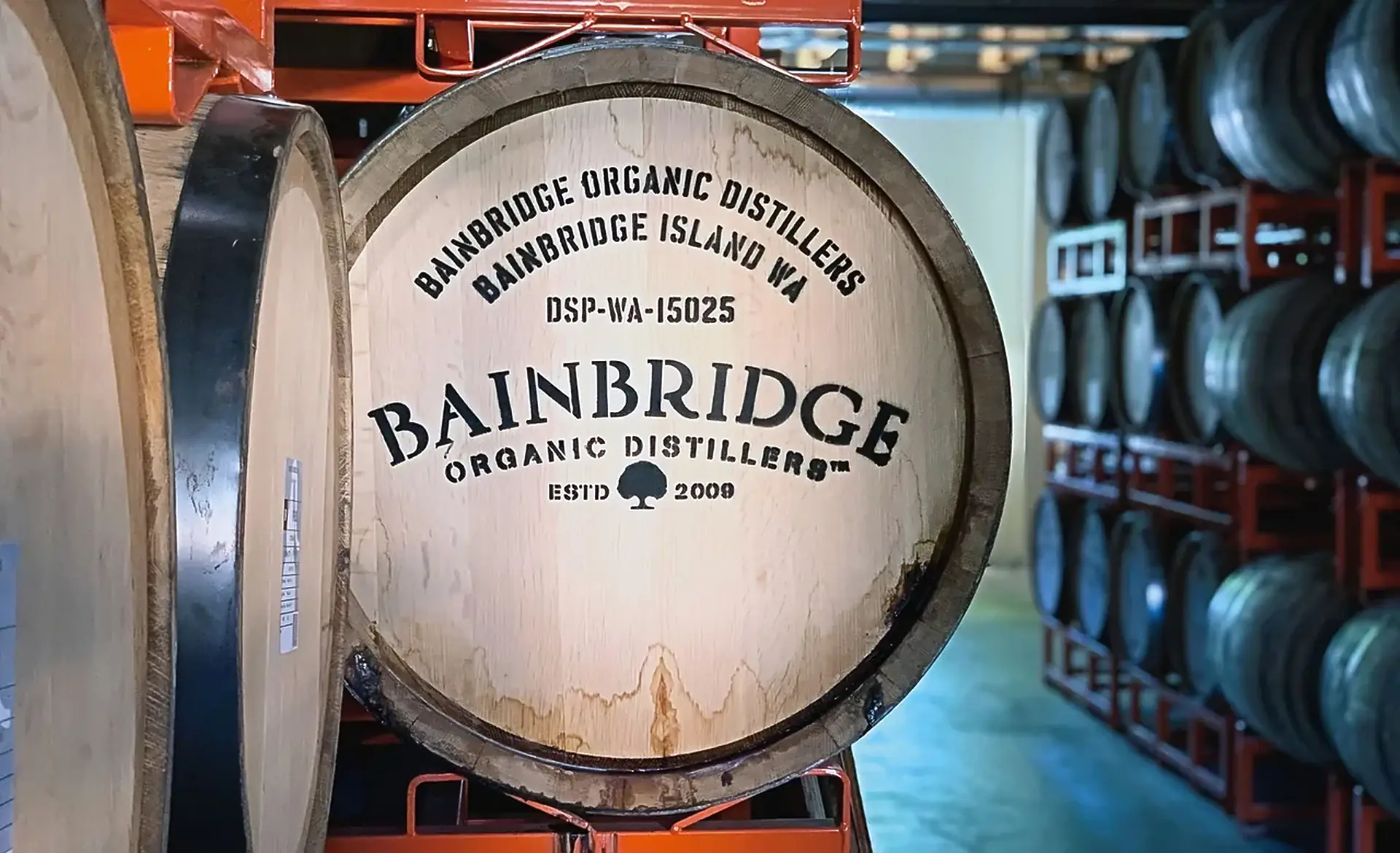 Barrel head stencilled with the Bainbridge name, Bainbridge Island WA and a federal plant number, with barrels racked behind