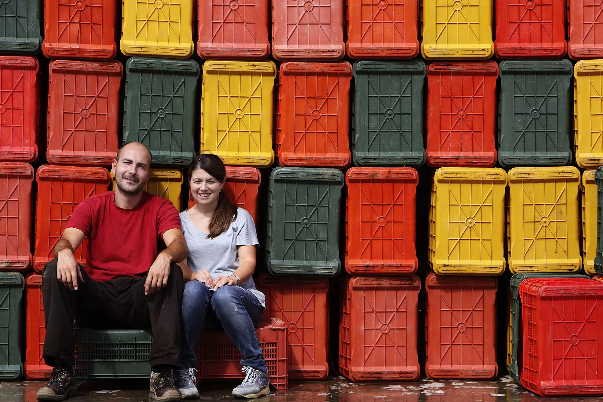 A man in a red T-shirt and a woman in a grey top sitting in front of a wall of stacked red, yellow and green plastic crates