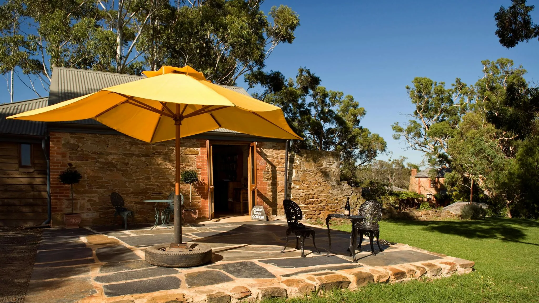 Stone cellar door with a yellow umbrella over a flagstone patio and gum trees behind, at Battle of Bosworth