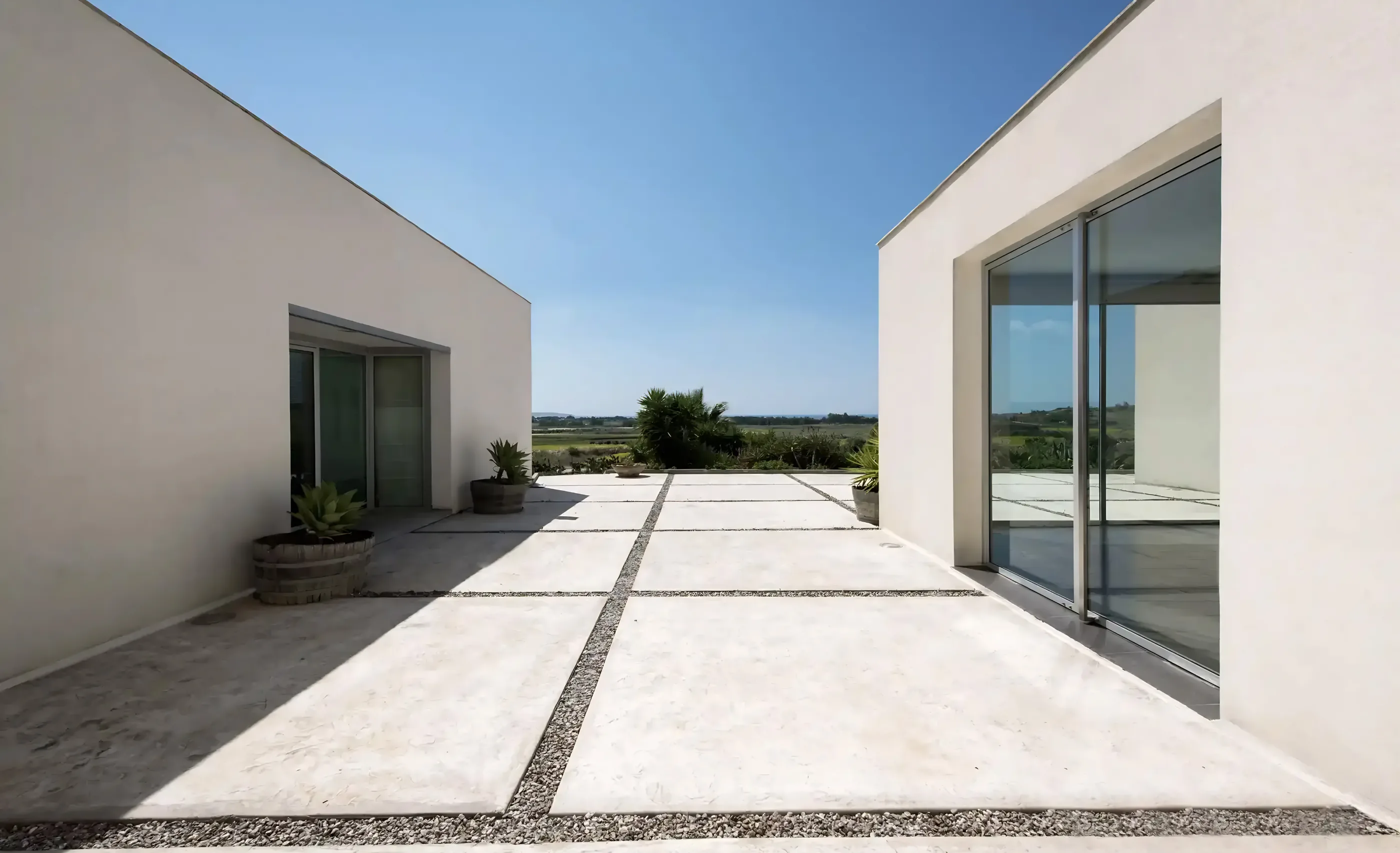 Two white, flat-roofed buildings around a paved courtyard with potted agaves, glass doors and green fields beyond