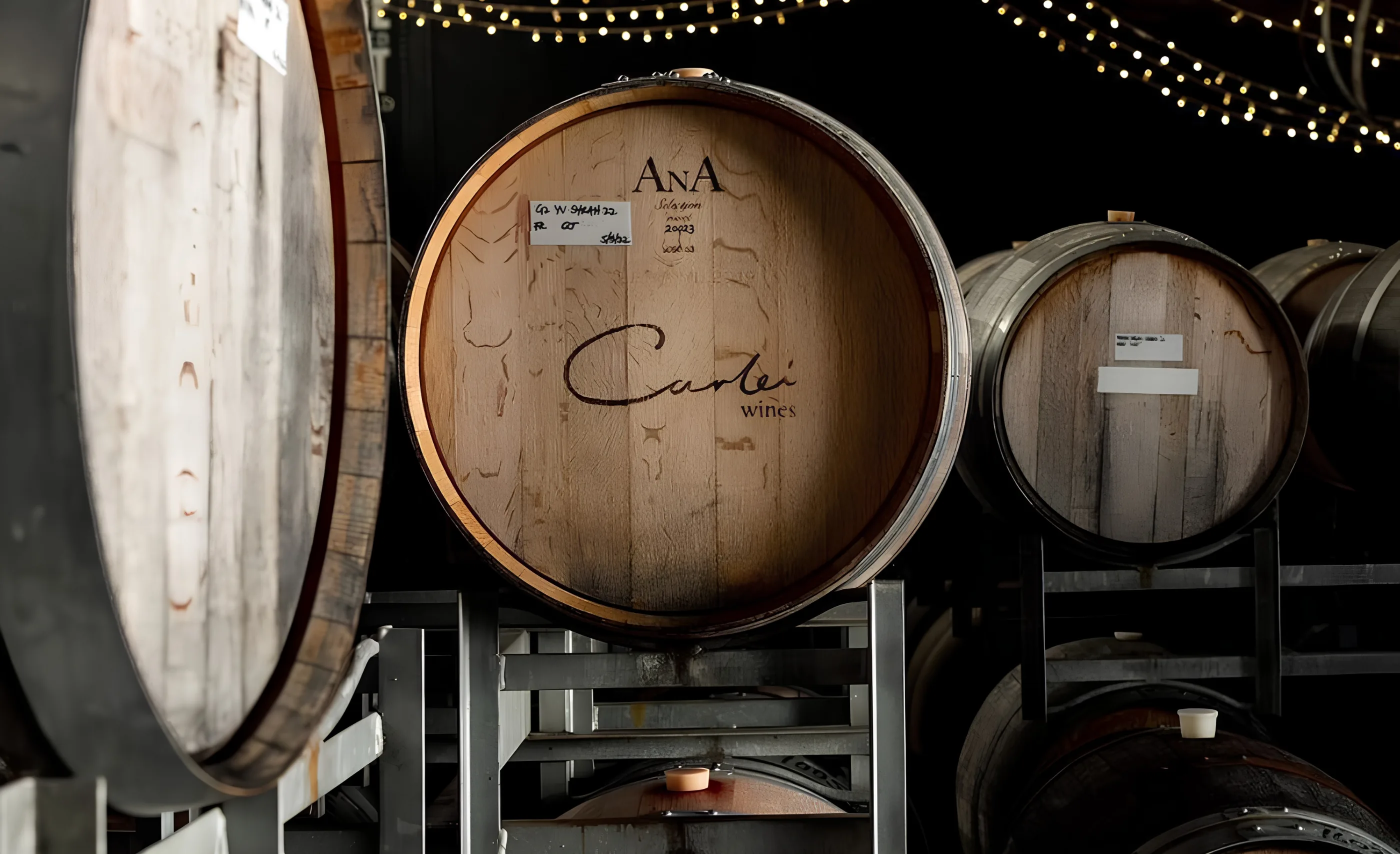 Oak barrel in a cellar under string lights, its head branded Carlei wines, with more barrels racked beside it