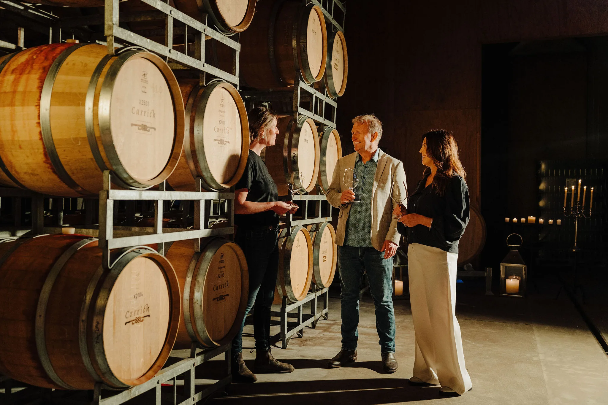 Three people holding wine glasses and talking beside racked barrels branded Carrick, with candles in the dim hall behind