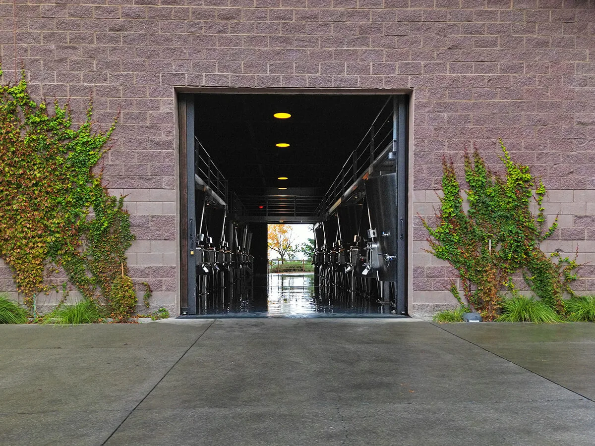 Open winery doorway in a block wall between ivy, with steel tanks either side and daylight at the far end