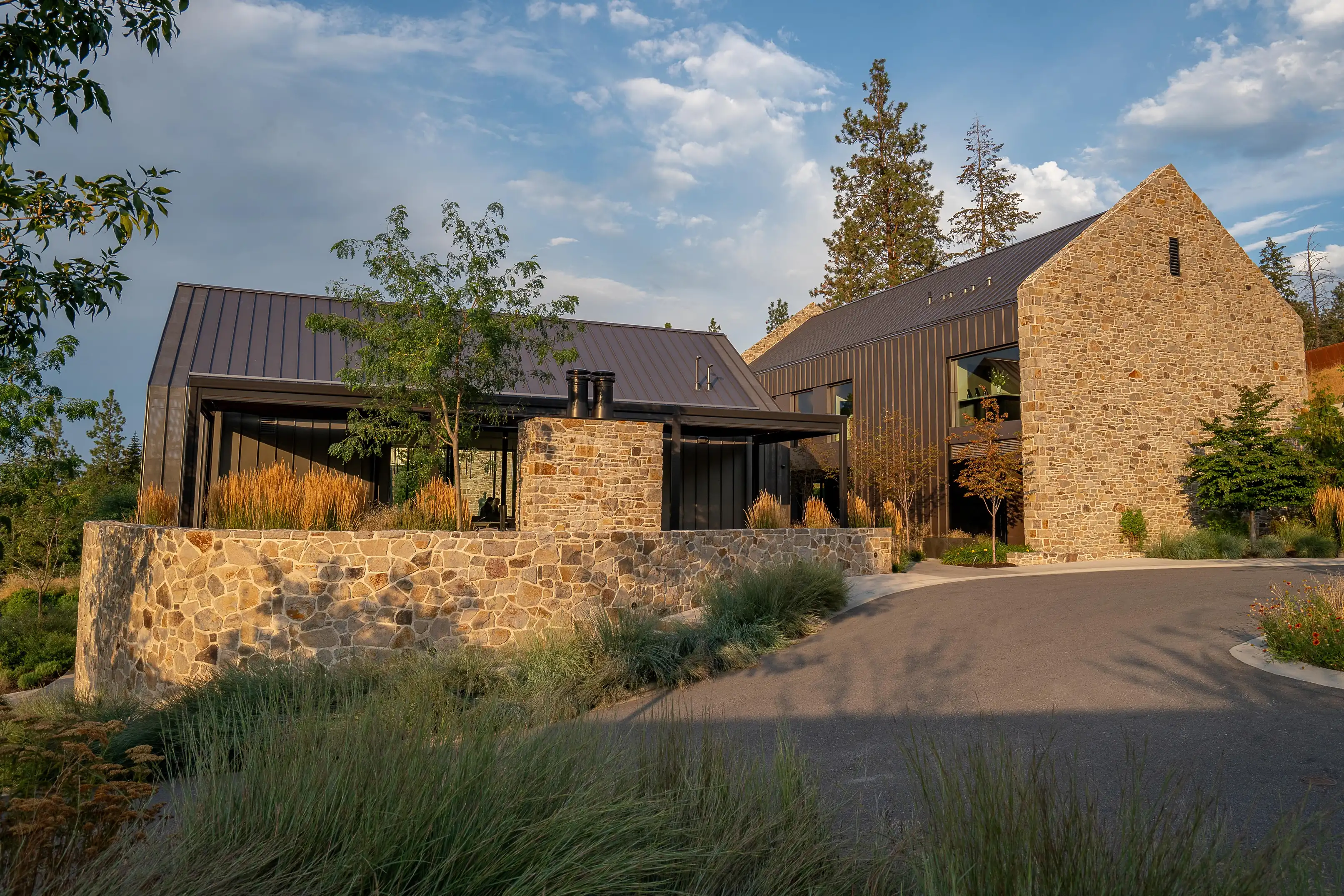Stone-gabled building and dark metal-clad wings with a curving drive and tall grasses, pines behind, in late sun