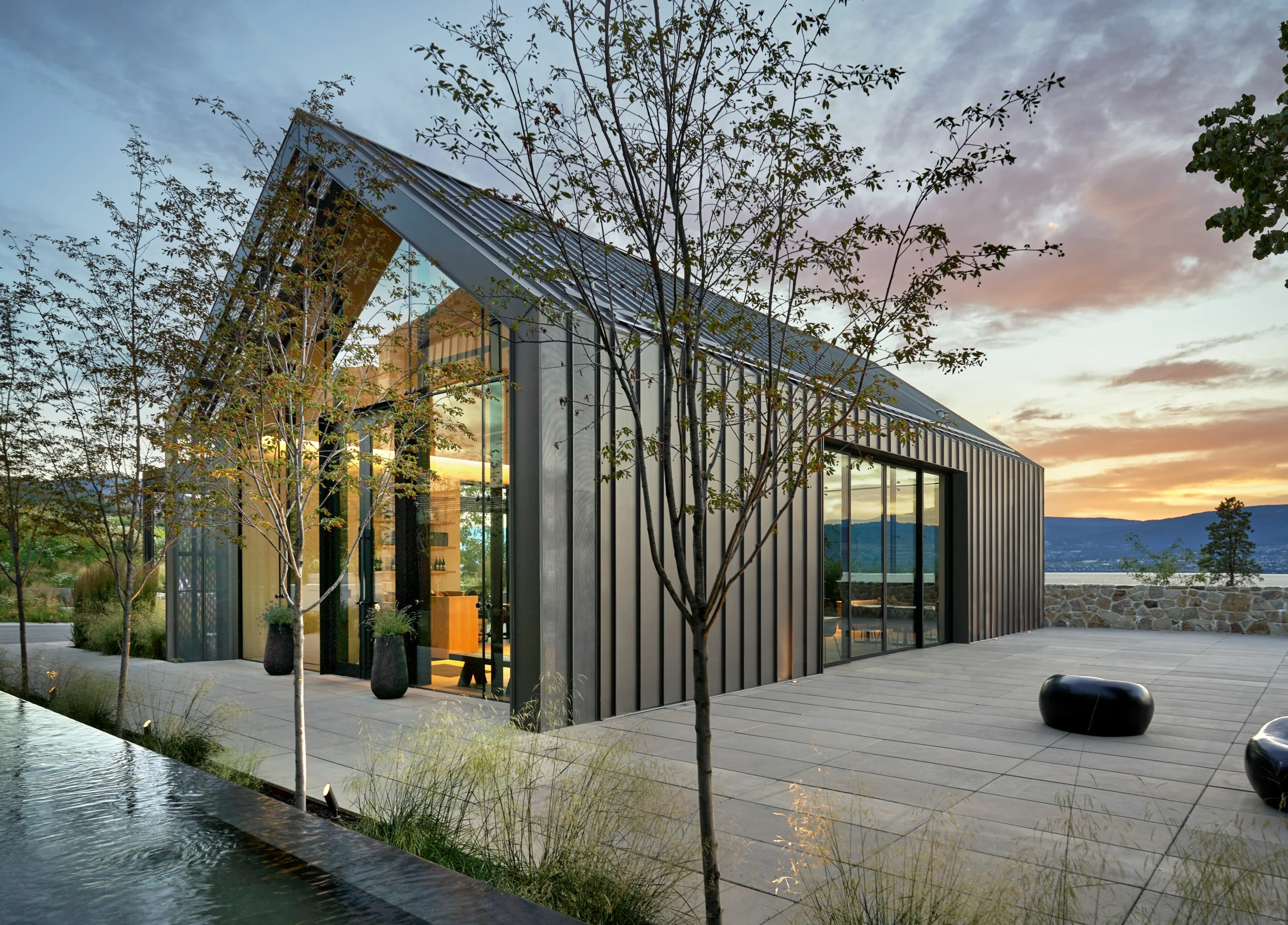 Dark metal-clad pavilion with a gabled glass front at dusk, young trees, a reflecting pool and distant hills under a pink sky