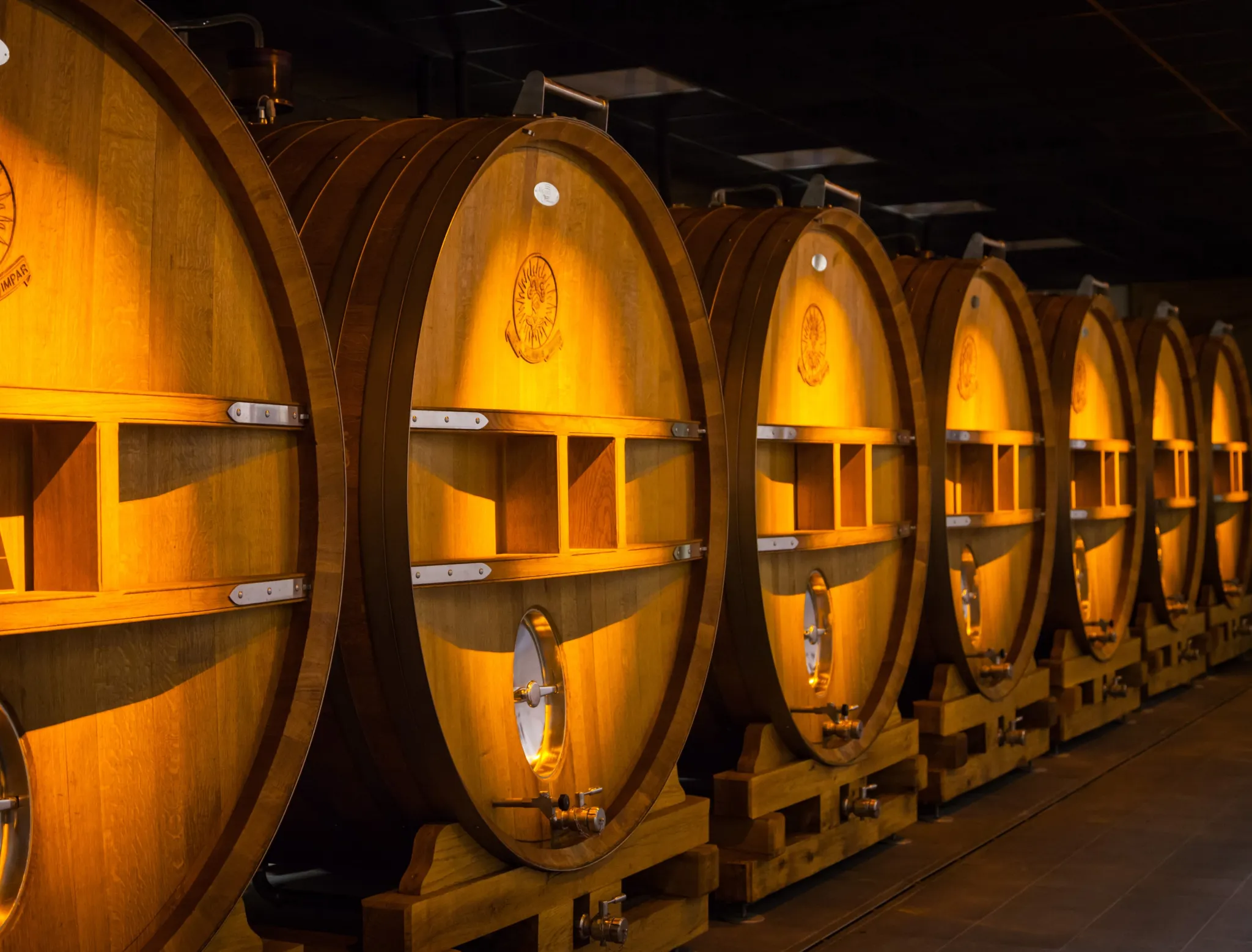 Row of large oak casks lit amber, each marked with a sun emblem, on wooden cradles in a dark hall