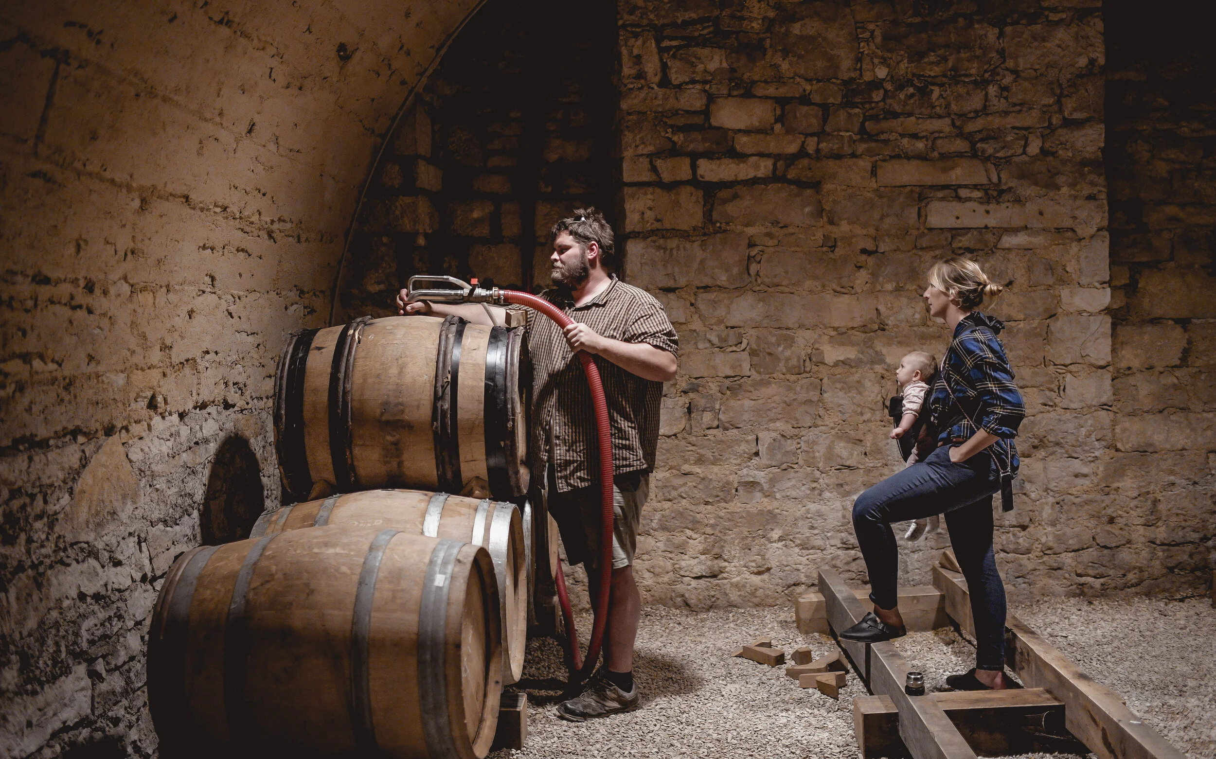 Bearded man works at stacked oak barrels with a red hose in a vaulted stone cellar while a woman carrying a baby looks on