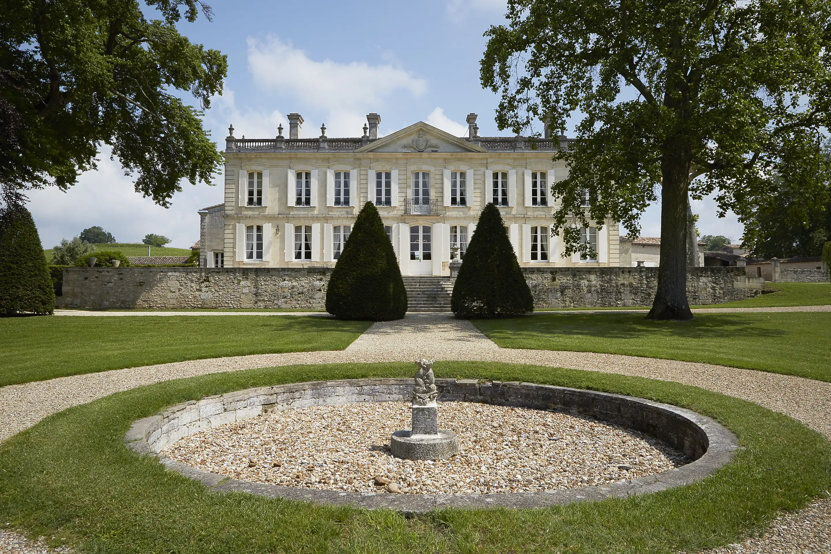 Pale stone château with a pediment and white shutters behind two cone-shaped topiary trees and a round gravel basin on a lawn
