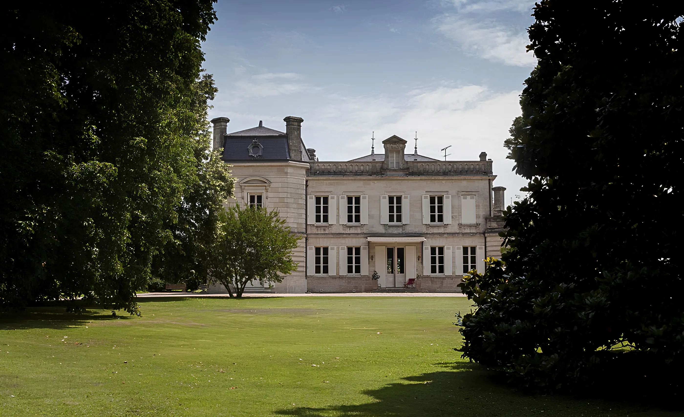 Pale stone château with a slate-roofed pavilion at left and white shutters, seen across a lawn between dark trees