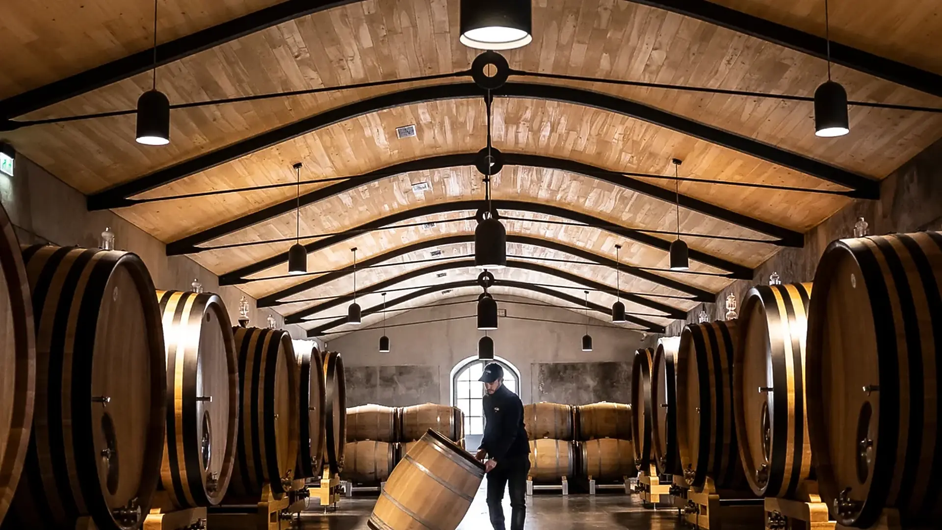 Man in a cap rolling an oak barrel along the aisle of a hall with an arched timber ceiling and large oak casks on both sides