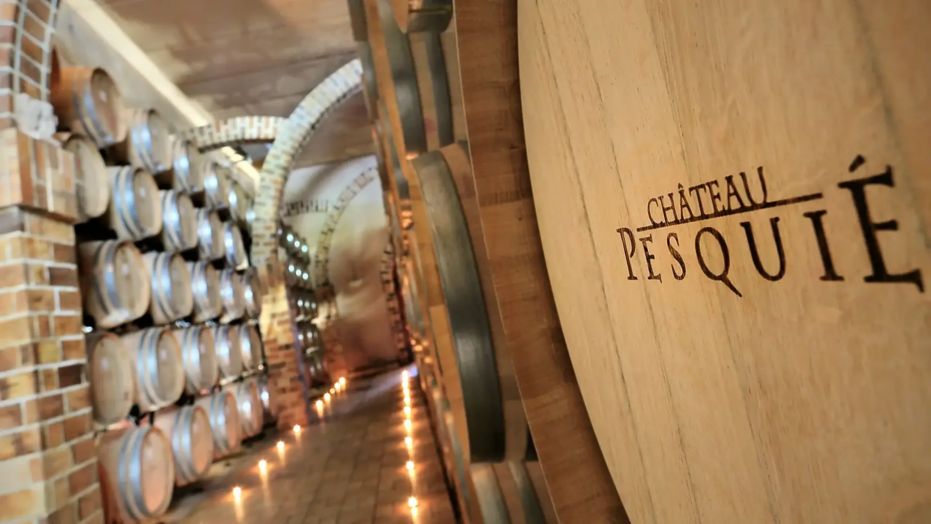 Oak cask end branded Château Pesquié beside stacked barrels in a brick-vaulted cellar, small lights along the floor
