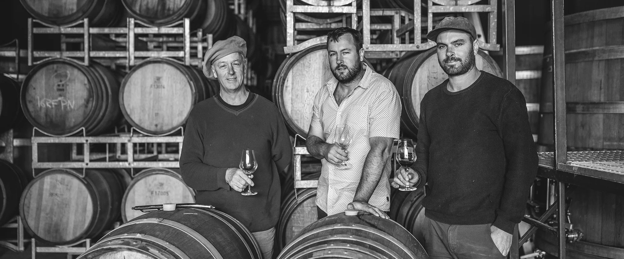 Three men holding wine glasses between stacked oak barrels in a barrel hall, in black and white
