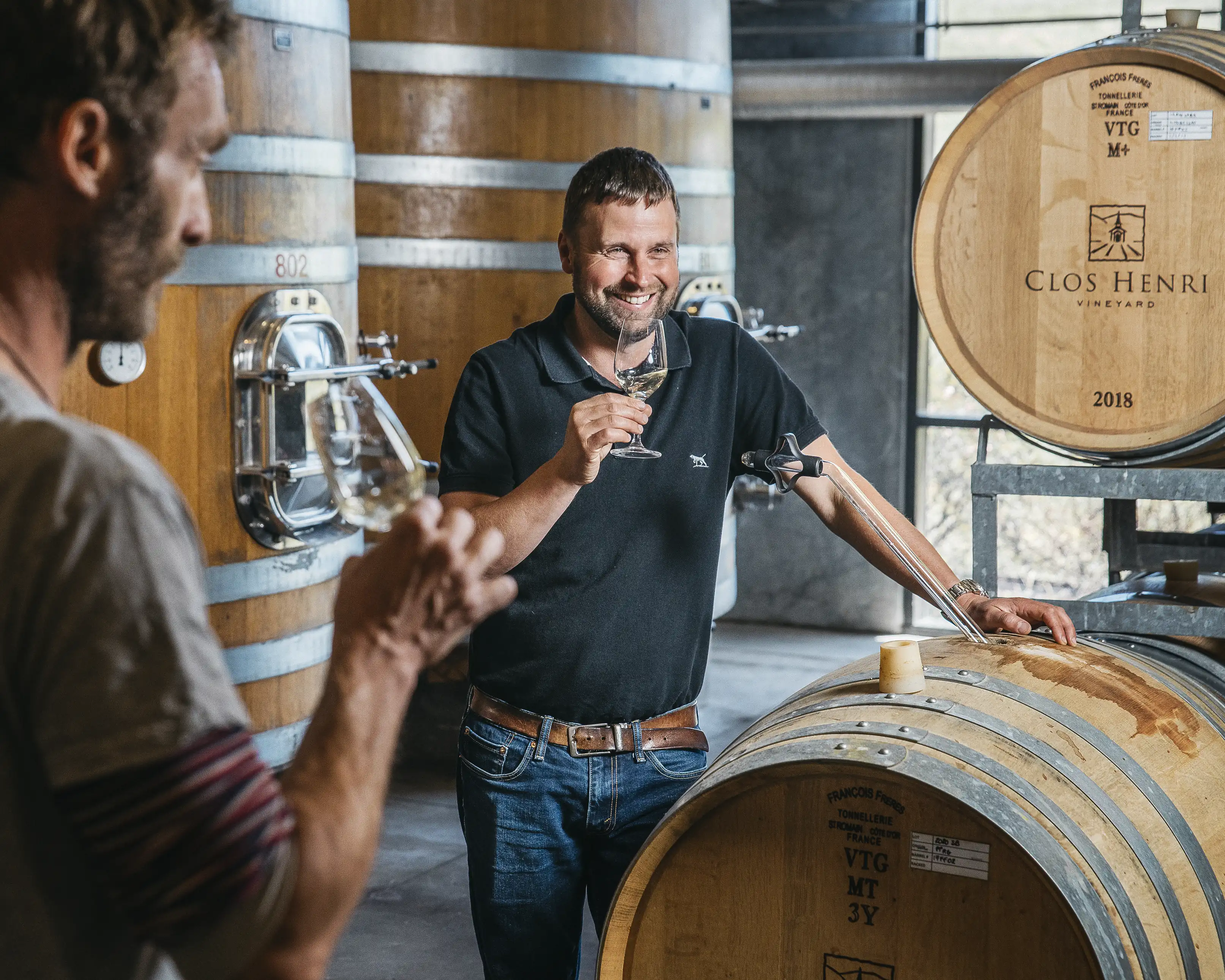 Two men tasting white wine among oak vats and barrels, one barrel head branded Clos Henri Vineyard