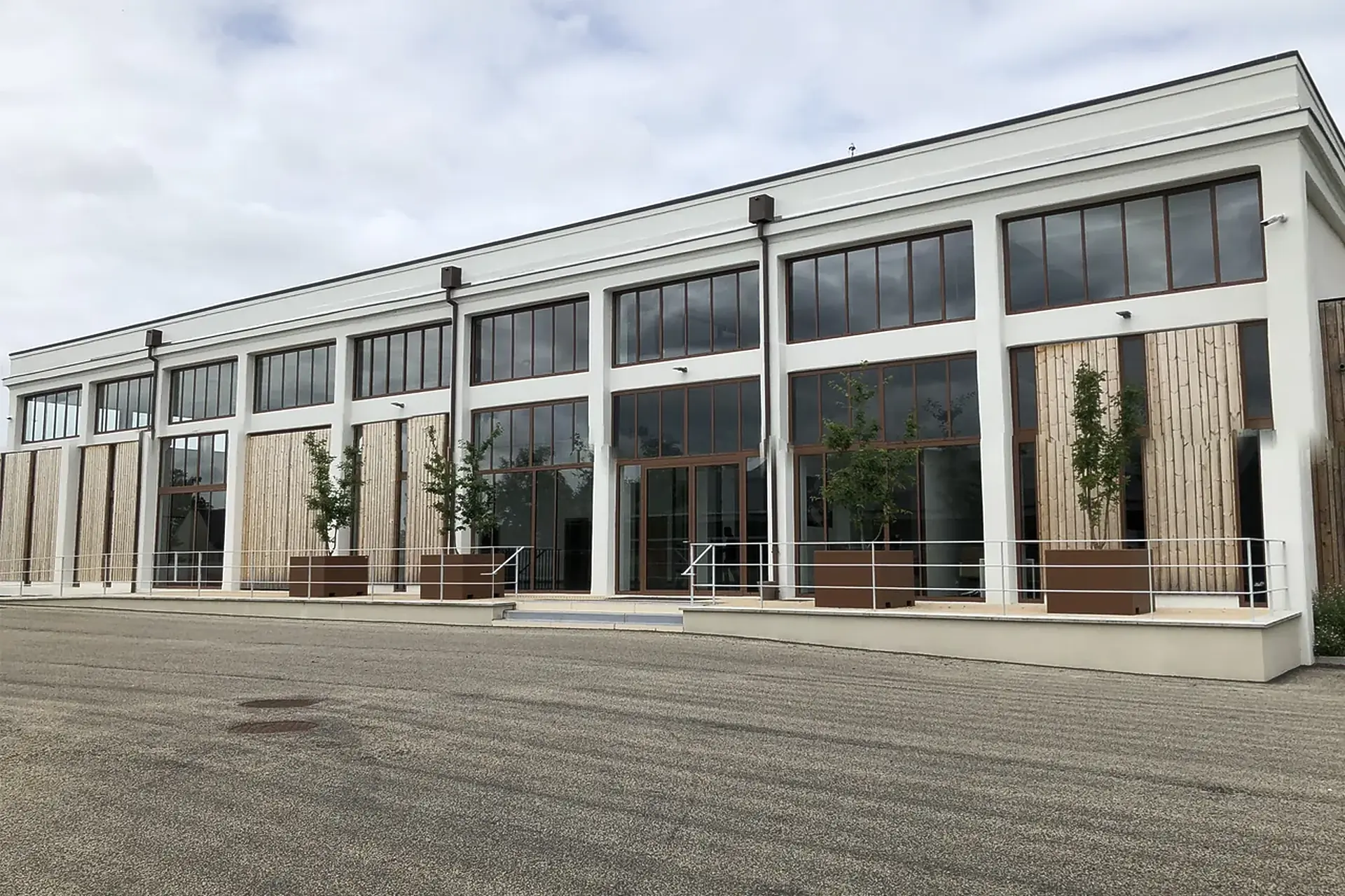 Long white two-storey building with tall brown-framed windows, timber-clad panels and small trees in planters, facing a gravel yard