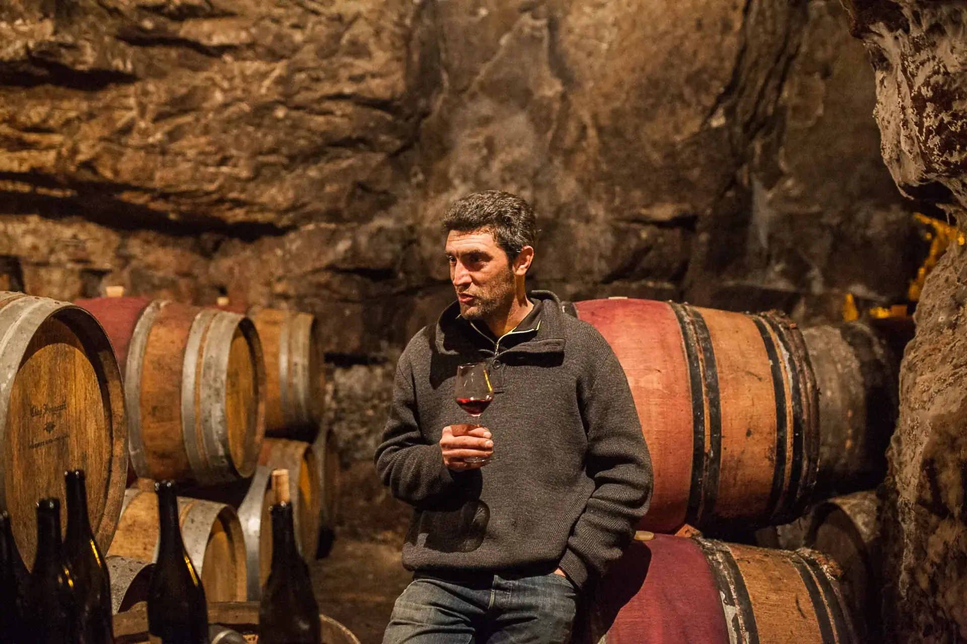 Man in a grey sweater holding a glass of red wine, leaning on barrels in a rough rock-cut cellar with bottles at lower left