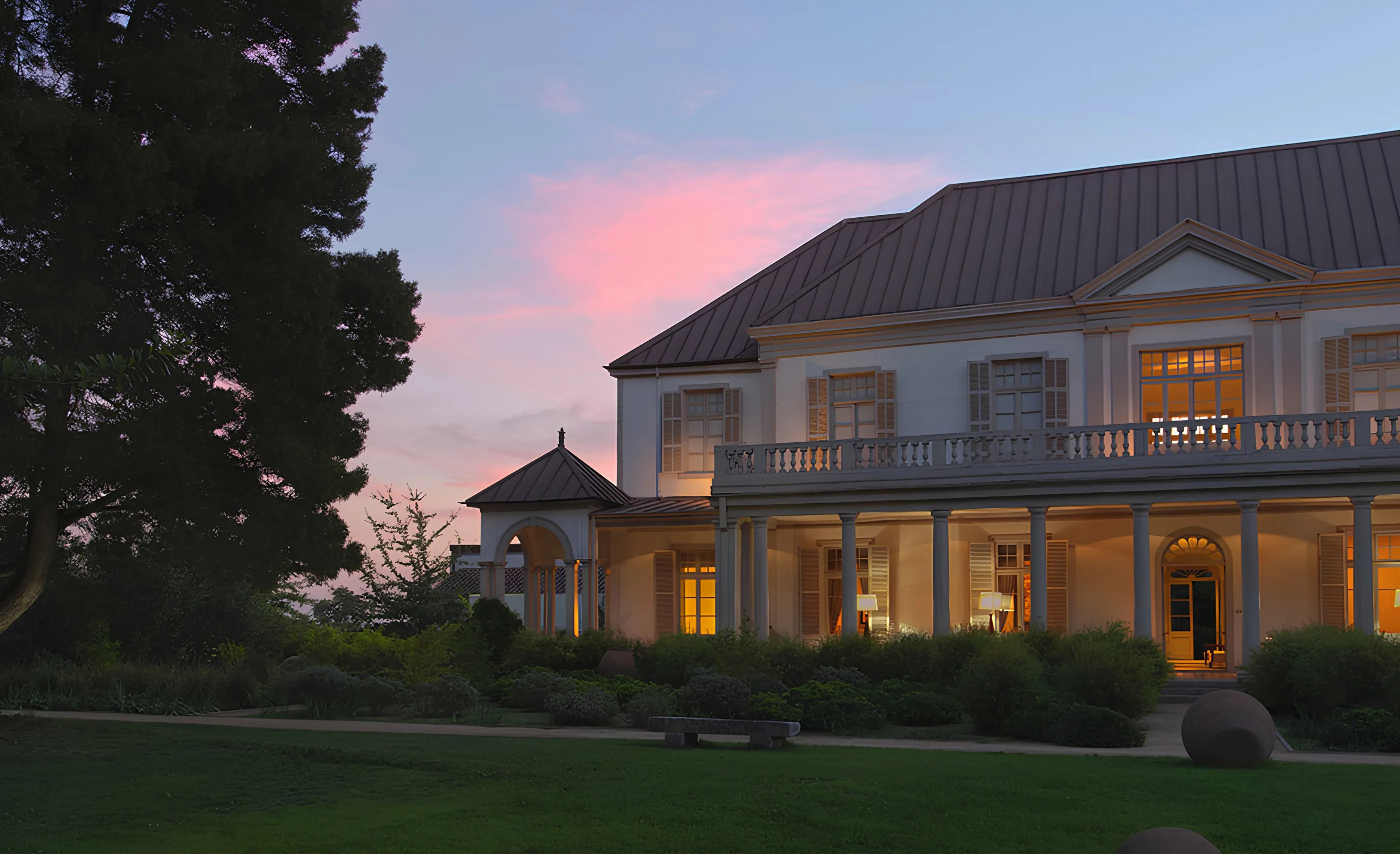 Cream-coloured house with a columned veranda, balcony and metal roof, windows lit at dusk under a pink sky, at Cono Sur