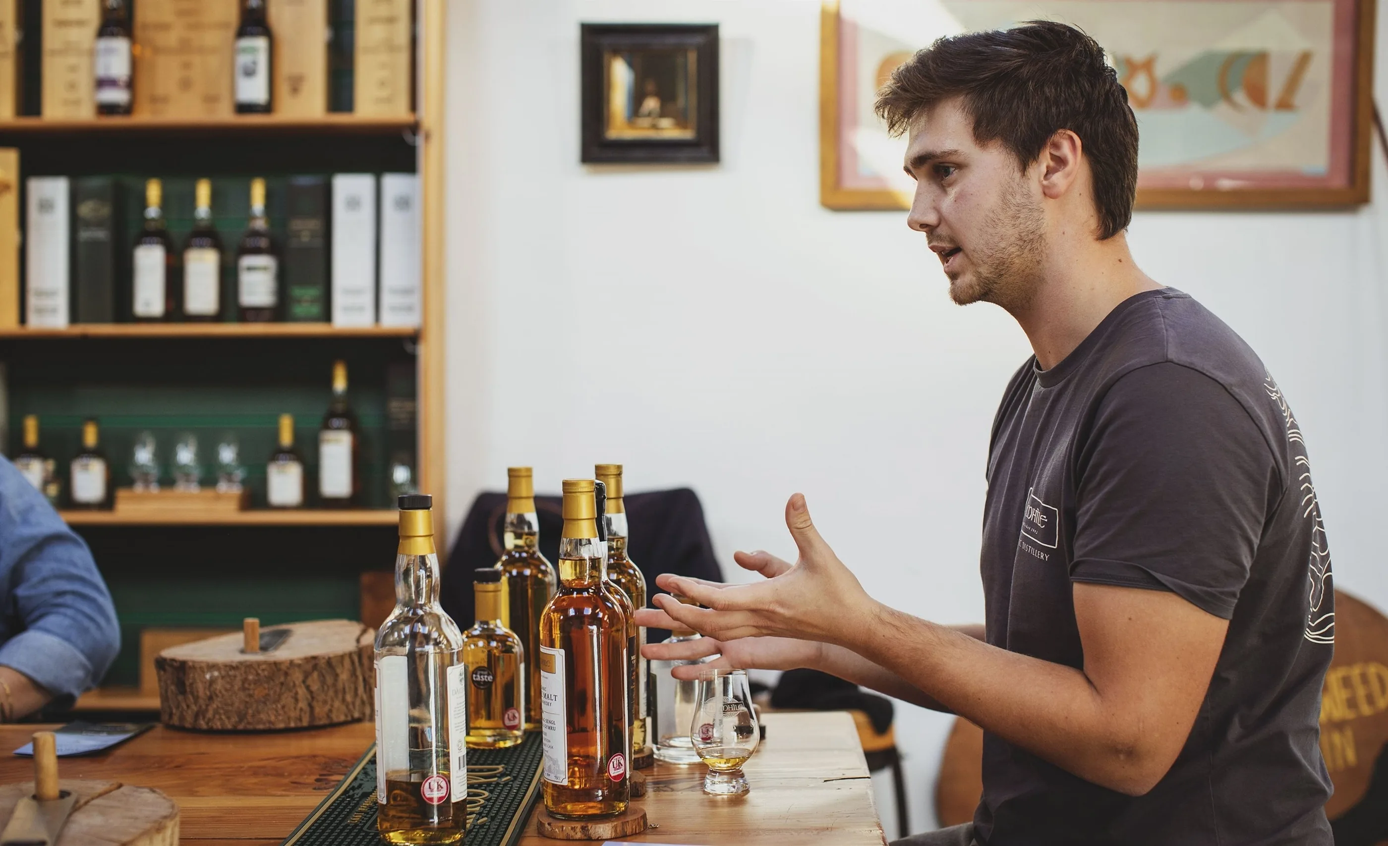A man in a grey t-shirt gestures as he talks, whisky bottles and a tasting glass on a wooden bar, shelves of boxed bottles behind