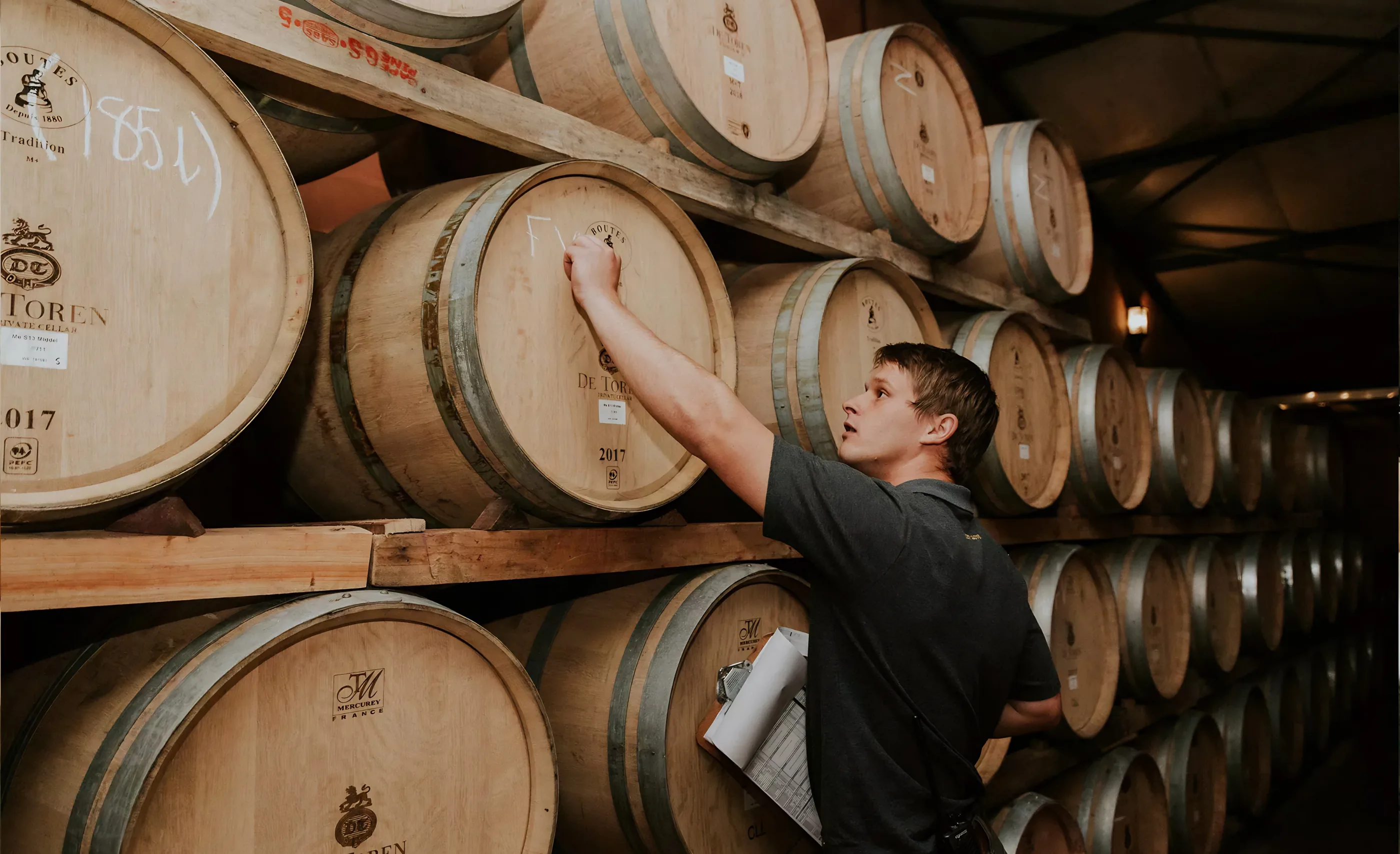 A man chalking a mark on a barrel in a stacked barrel cellar, the barrels branded De Toren
