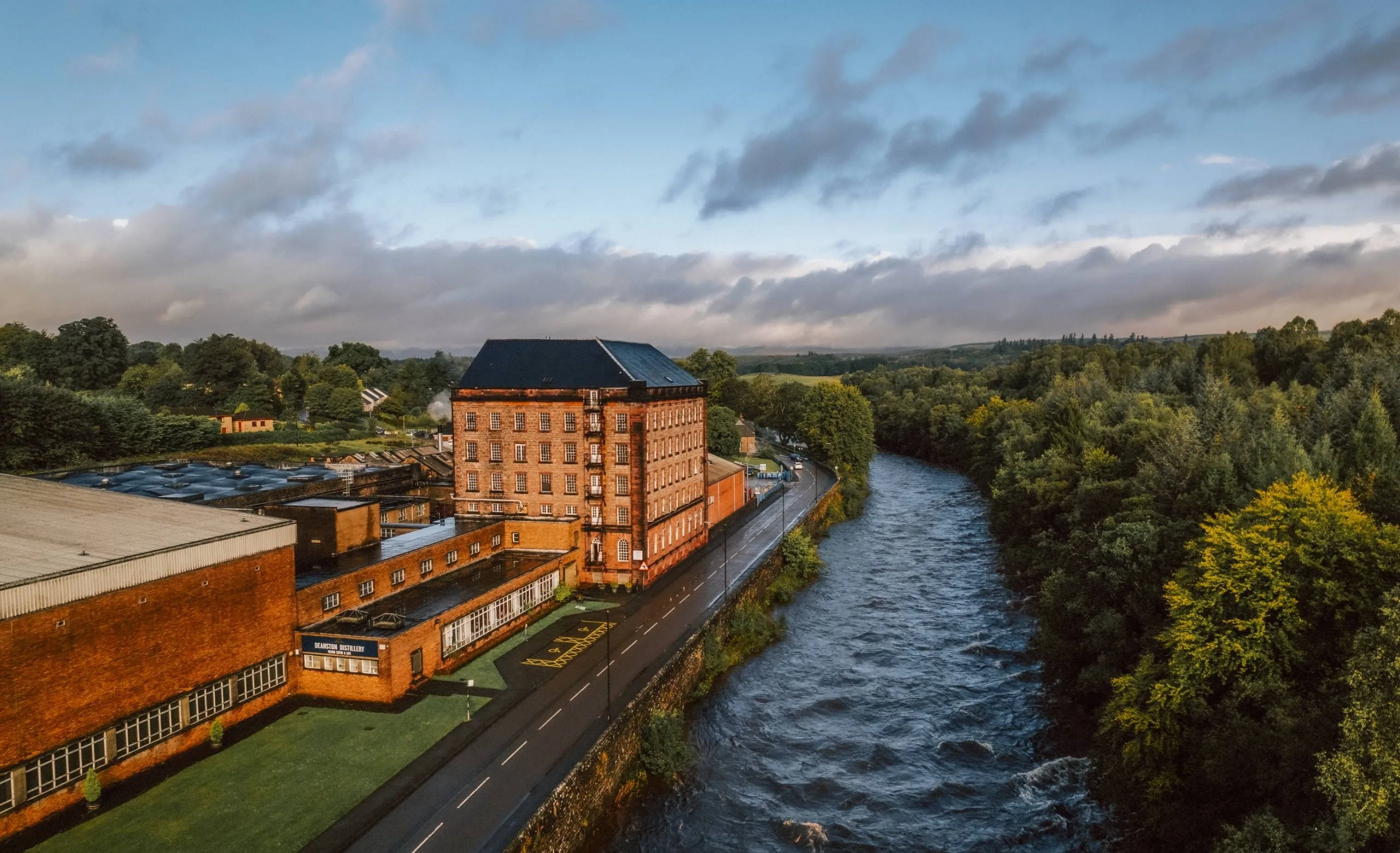 Aerial view of Deanston Distillery's red-brick buildings beside a fast-flowing river and road, with woods along the far bank