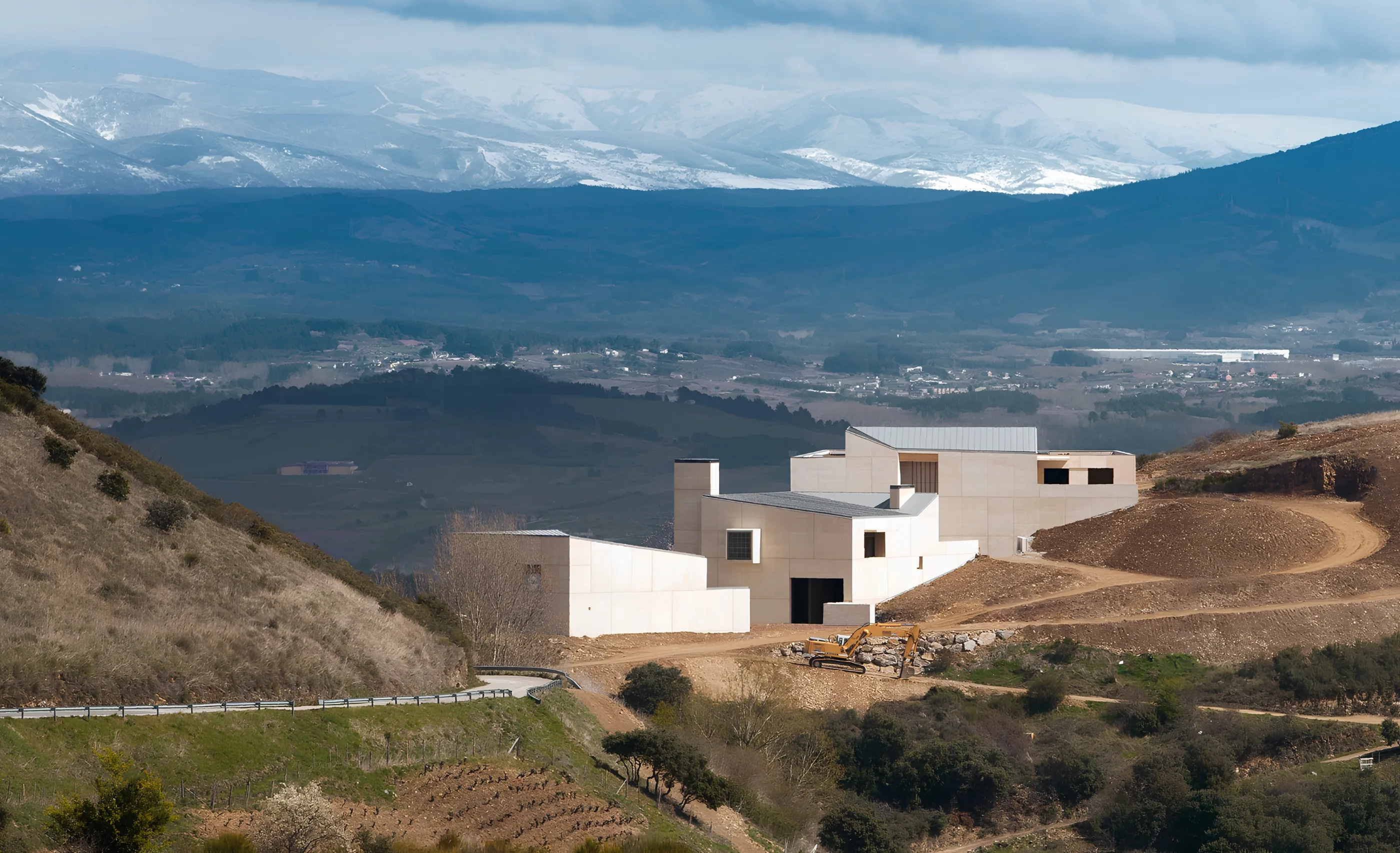 A white modern building on a bare hillside with an excavator beside it, a valley below and snow-covered mountains behind