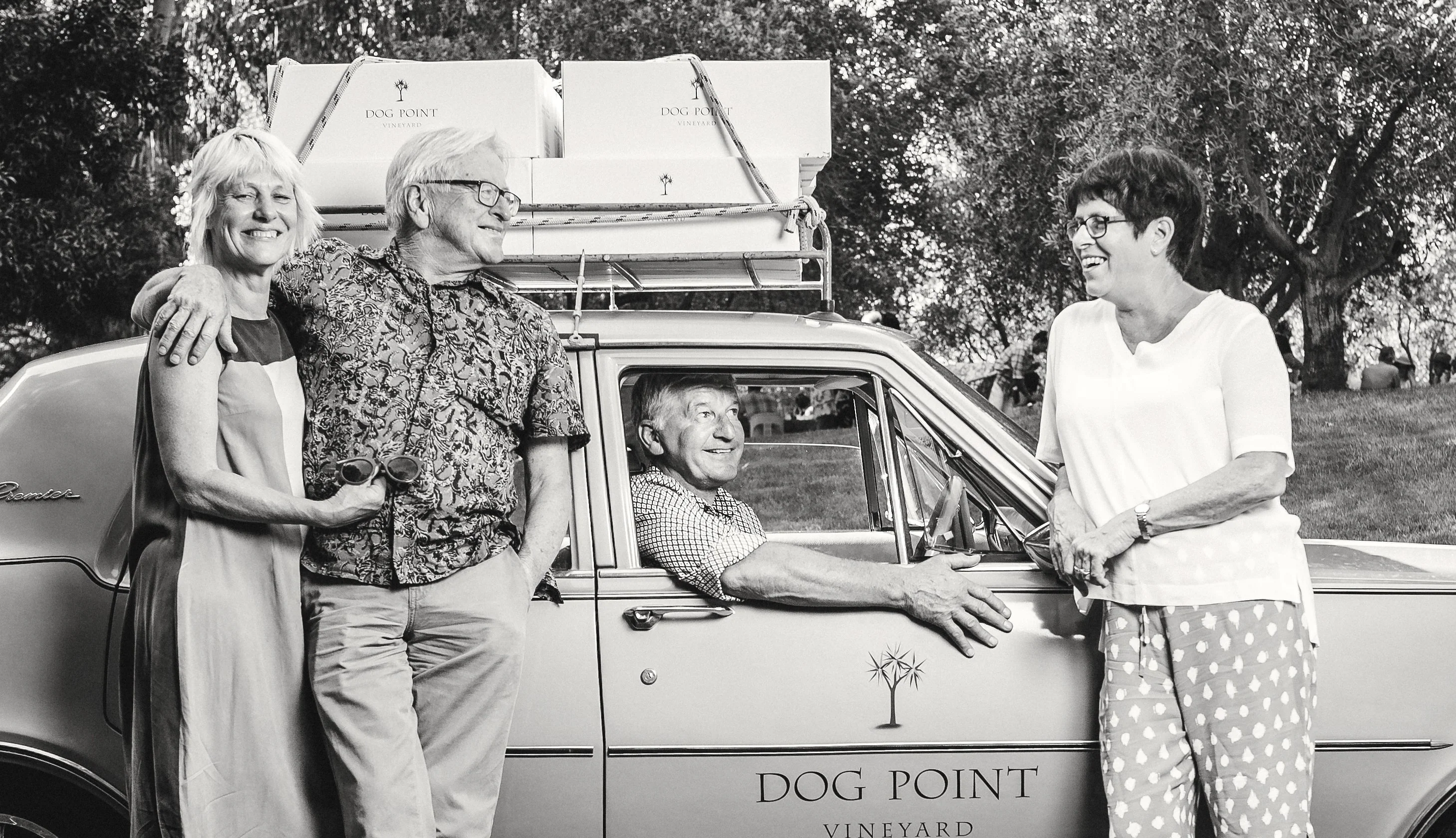 Two women and two men around a vintage car marked Dog Point Vineyard, with cartons roped to its roof rack, in black and white