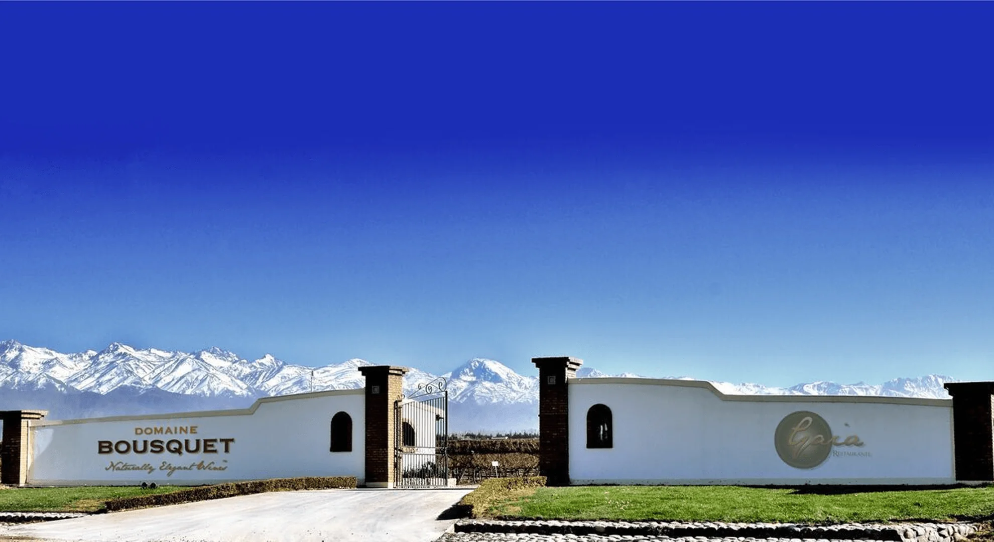 A white wall with a brick-pillared gate and a Domaine Bousquet sign, snow-capped mountains behind a deep blue sky