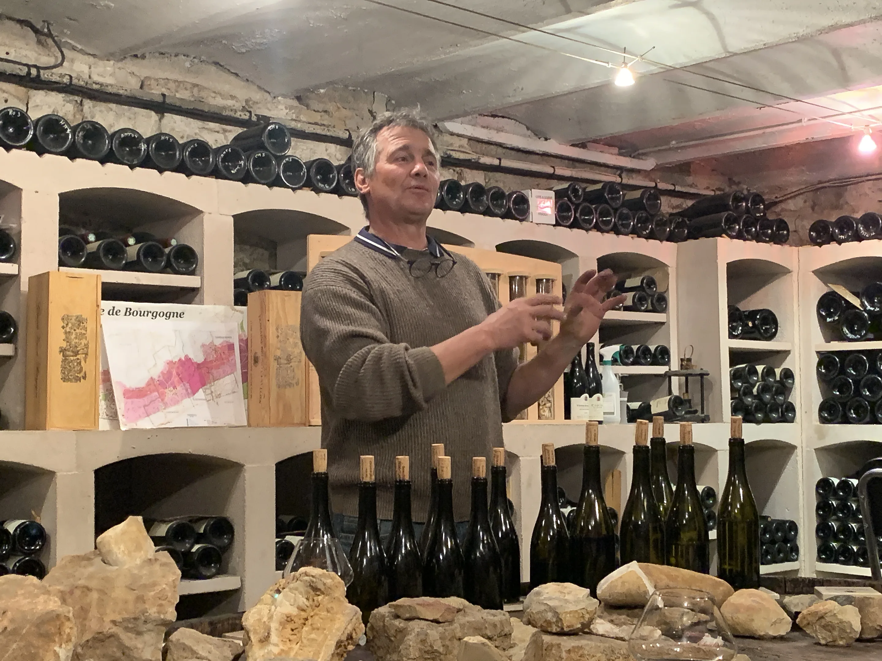 A man in a brown jumper talks with raised hands in Domaine Bruno Clavelier's tasting room, limestone rocks in front