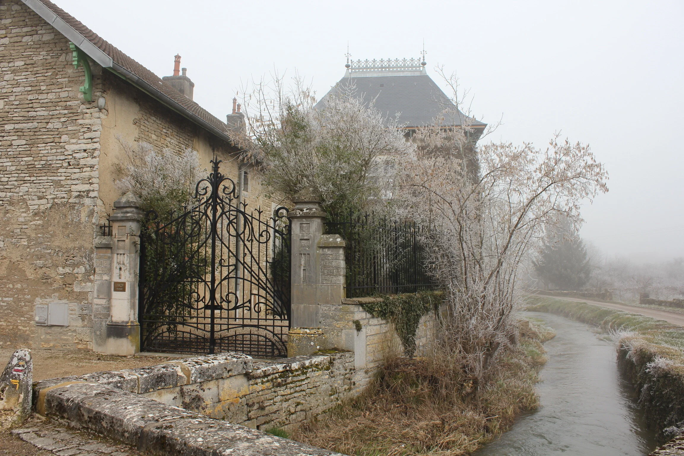 A stone building with a wrought-iron gate and pillars, a slate-roofed house behind frosted trees, and a narrow stream in mist