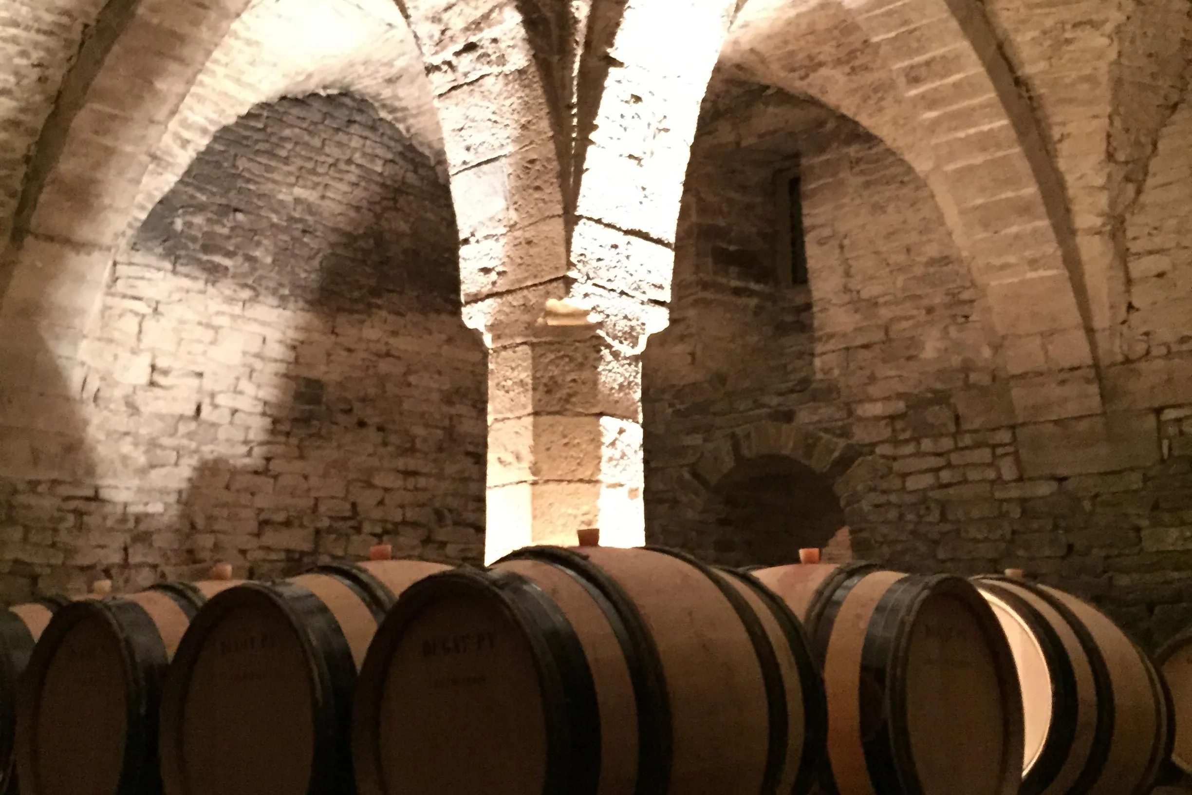 A vaulted stone cellar with ribbed arches springing from a lit central pillar, casks stencilled Dugat-Py in a row below
