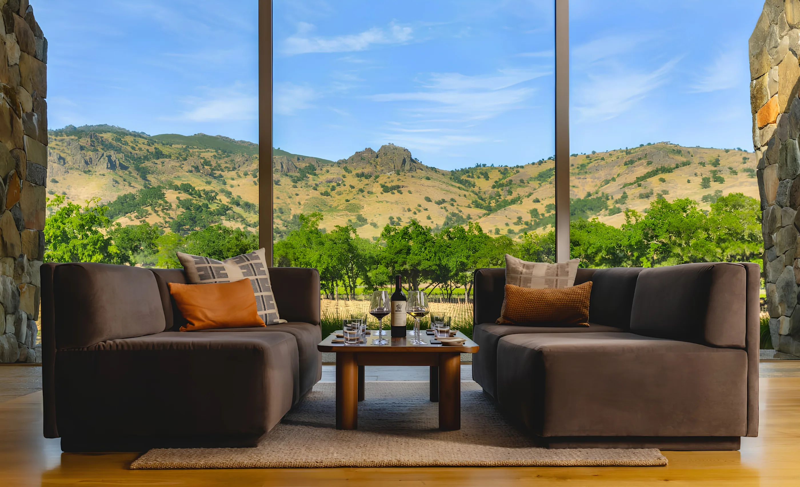 Two brown sofas around a low table with a bottle of red wine and glasses, before floor-to-ceiling windows onto dry golden hills