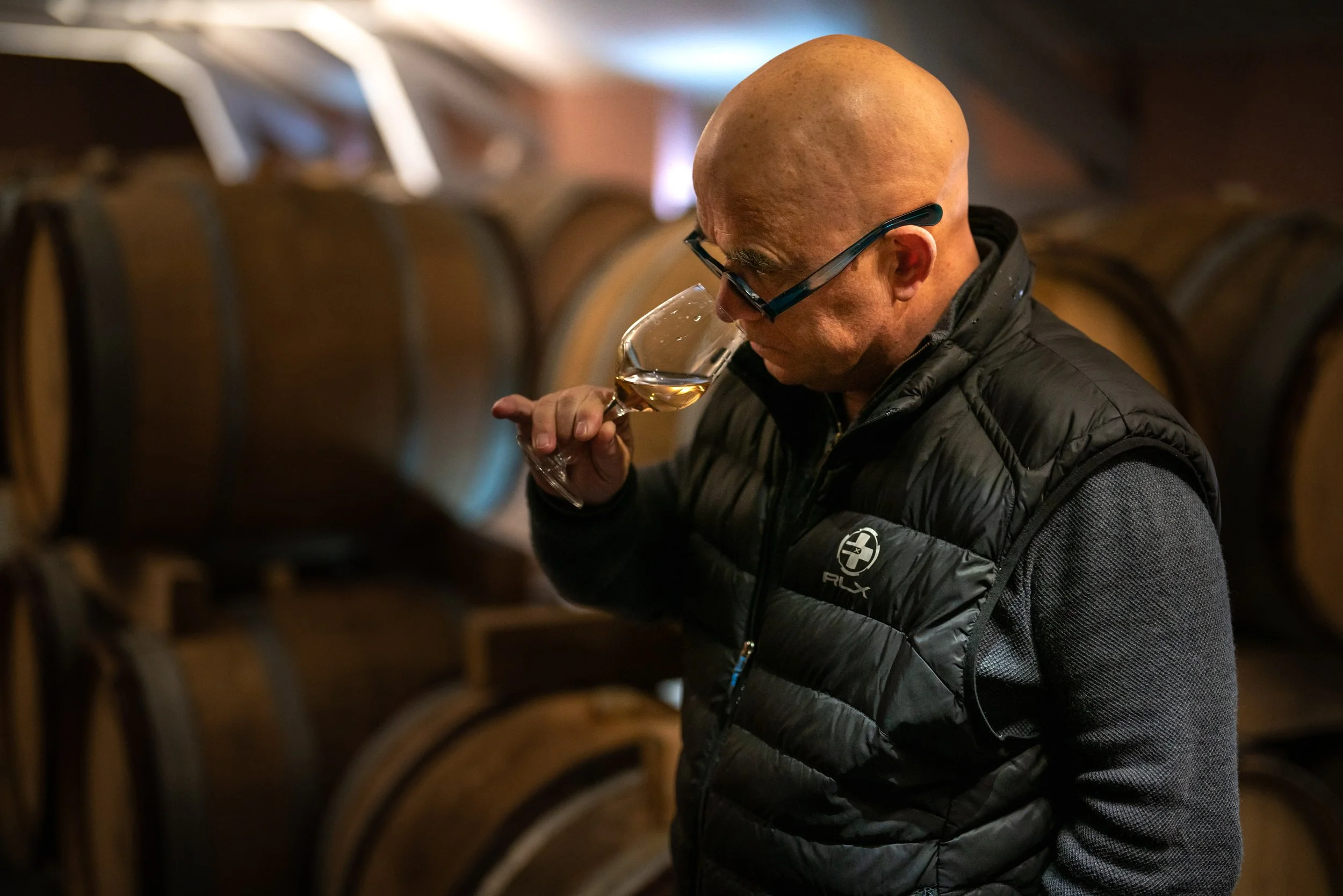 A bald man in glasses and a black quilted vest smells a glass of pale gold wine among stacked oak barrels