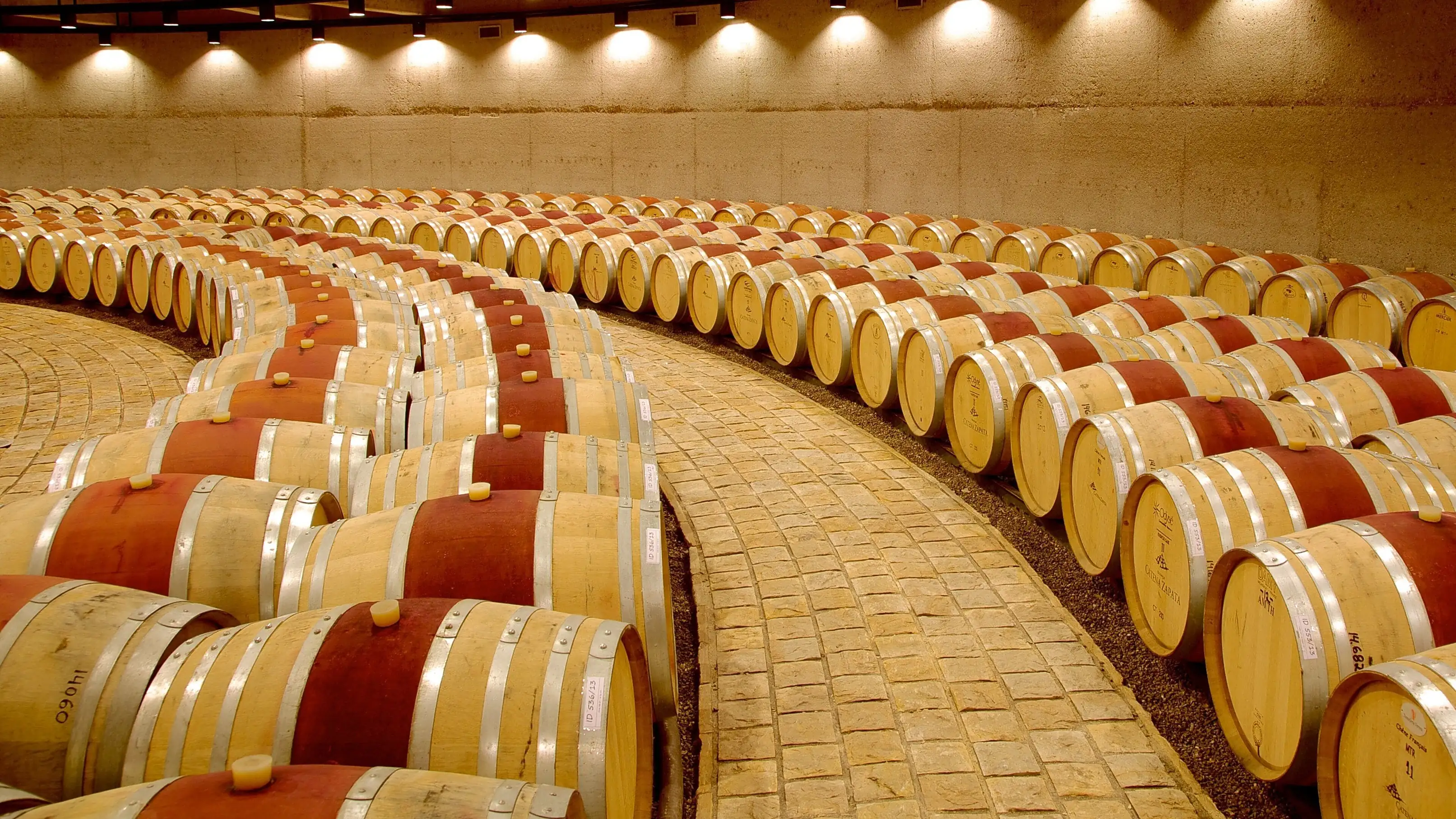 Oak barrels laid in curved rows on both sides of a cobbled path in a round cellar lit by spotlights