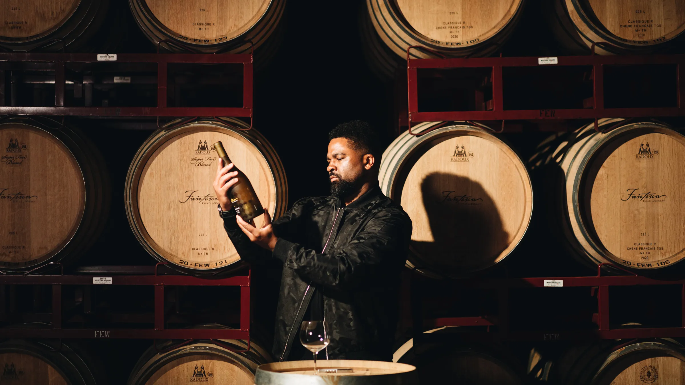 A man in a black jacket holds a bottle up to study it in the barrel room, with Fantesca barrels racked behind him