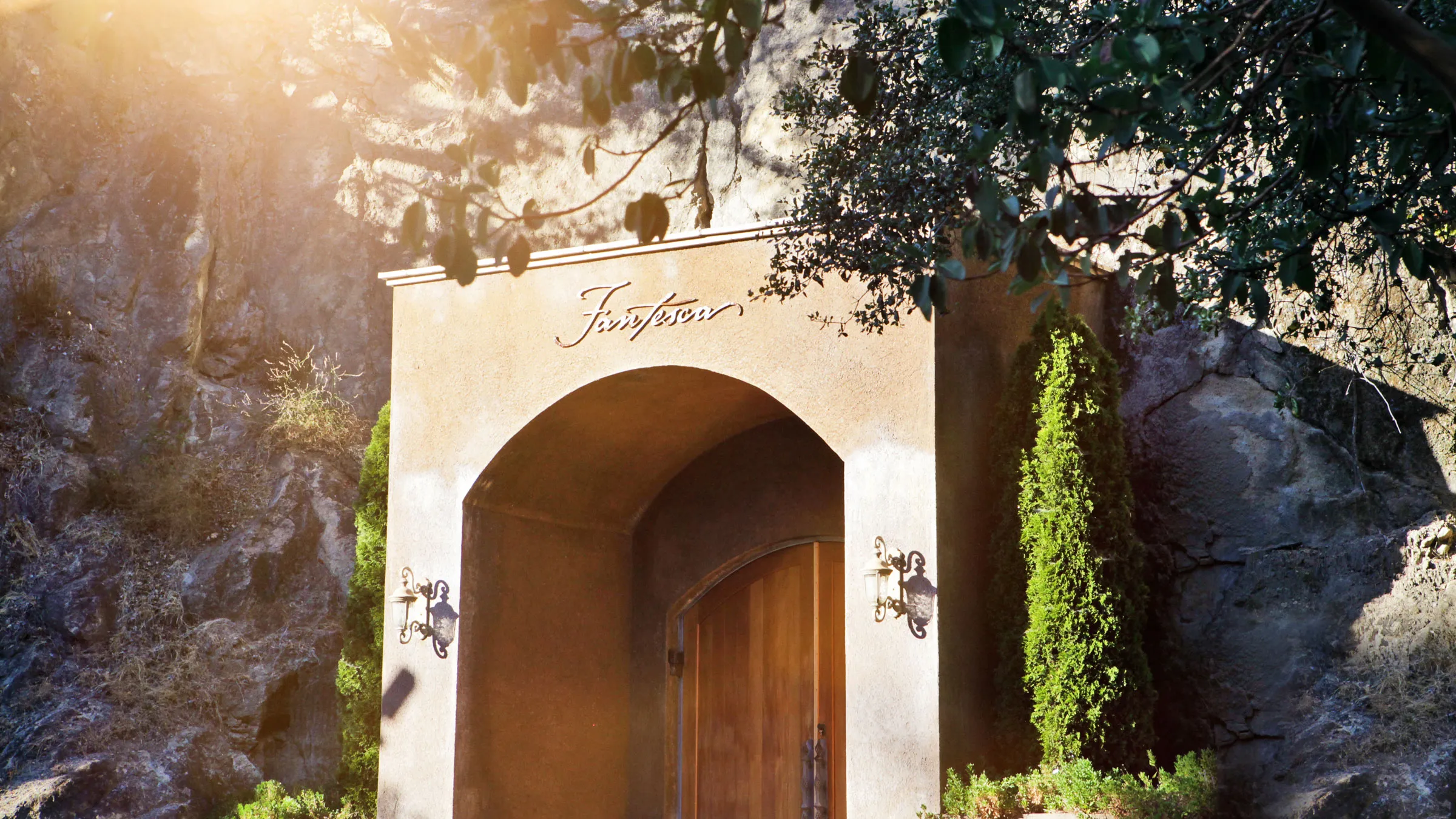 Arched cave entrance with the Fantesca name above a wooden door, set into a rock face under trees in low sun