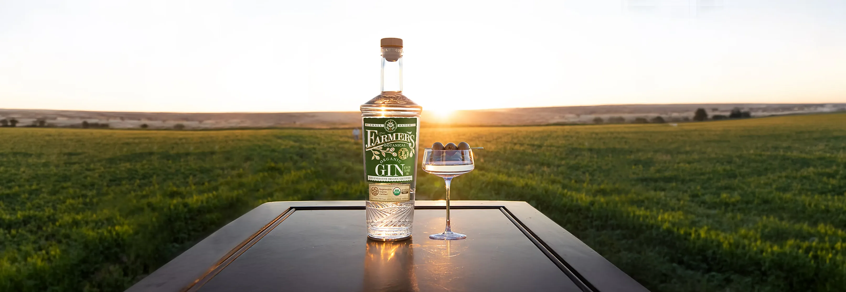A bottle of Farmer's Botanical gin beside a coupe glass on a dark table, with a green field and a low sun behind